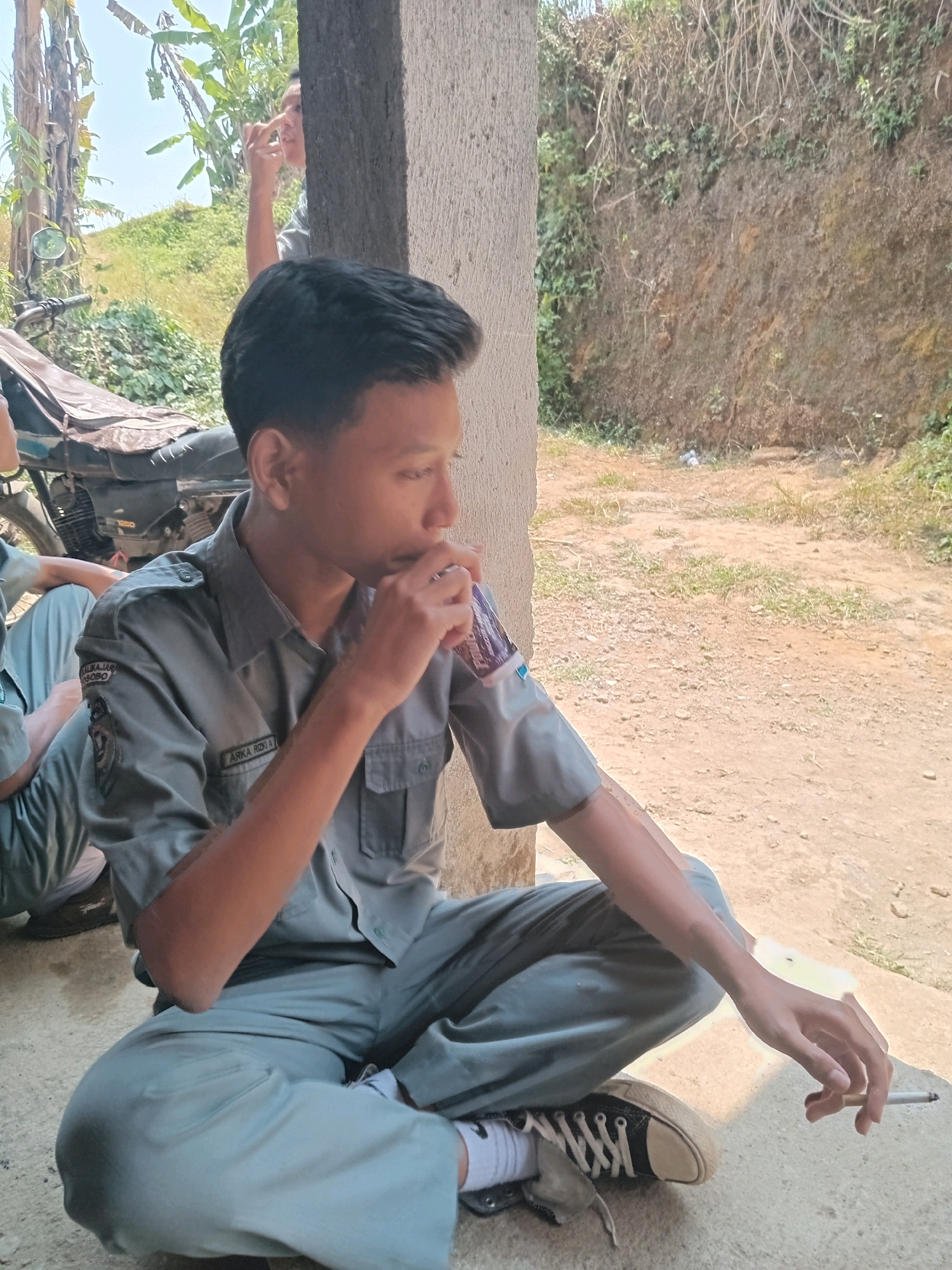 A young person in a light gray uniform is sitting cross-legged on the ground, with a cigarette in their right hand and a drink in their left. They are wearing a light gray collared shirt with patches on the sleeves and light gray pants, along with white socks and black and tan sneakers. The person is looking down and to their right. In the background, another person is partially visible behind a concrete pillar, with their hand near their face. A motorcycle with a brown seat cover is parked to the left. The environment appears to be outdoors, with a dirt ground, dry grass, and a steep, vegetated hillside in the background. The presence of a concrete pillar and the overall setting suggest this location is likely within the village of Kalikajar, Wonosobo Regency, Indonesia, perhaps near a school or community area, given the uniform.
