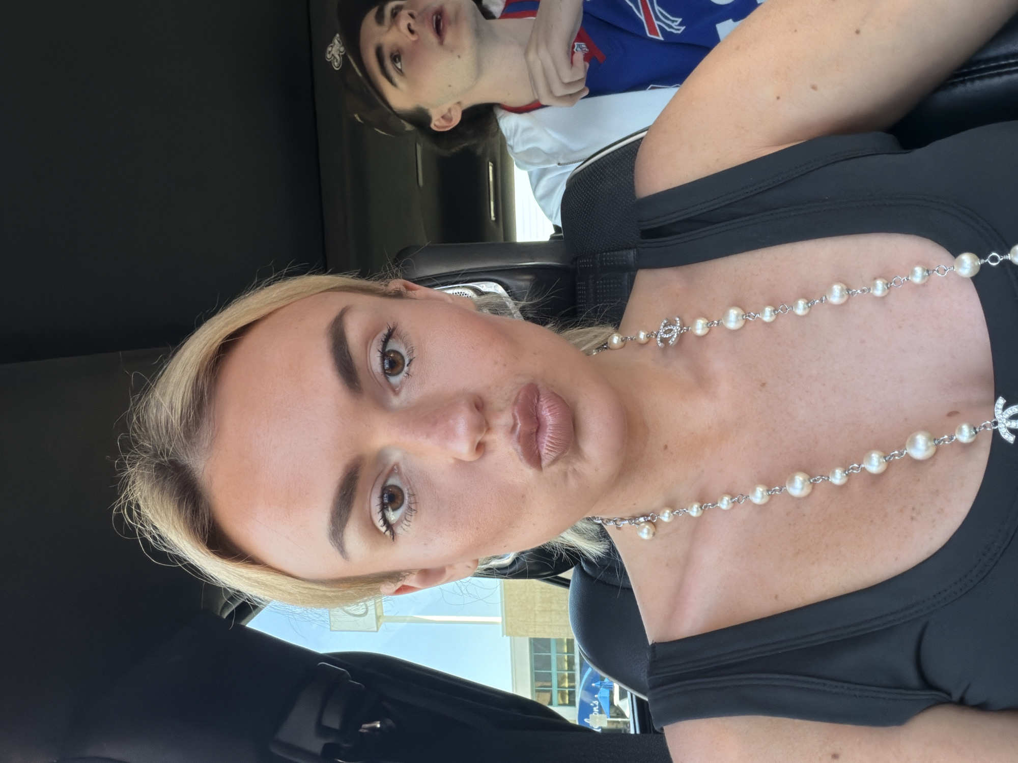 The image presents a selfie-style view inside a car, showing a woman with blonde hair and brown eyes positioned in the foreground. She is wearing a black top and a pearl necklace with a Chanel logo. In the background, partially visible, is a young man in a baseball cap and a sports jersey that includes the colors red, white, and blue.
Based on the visible outside background through the car windows, which includes a building with windows and a blue sign, it's likely the location is somewhere in an urban or suburban area in the city of Los Angeles, United States of America.