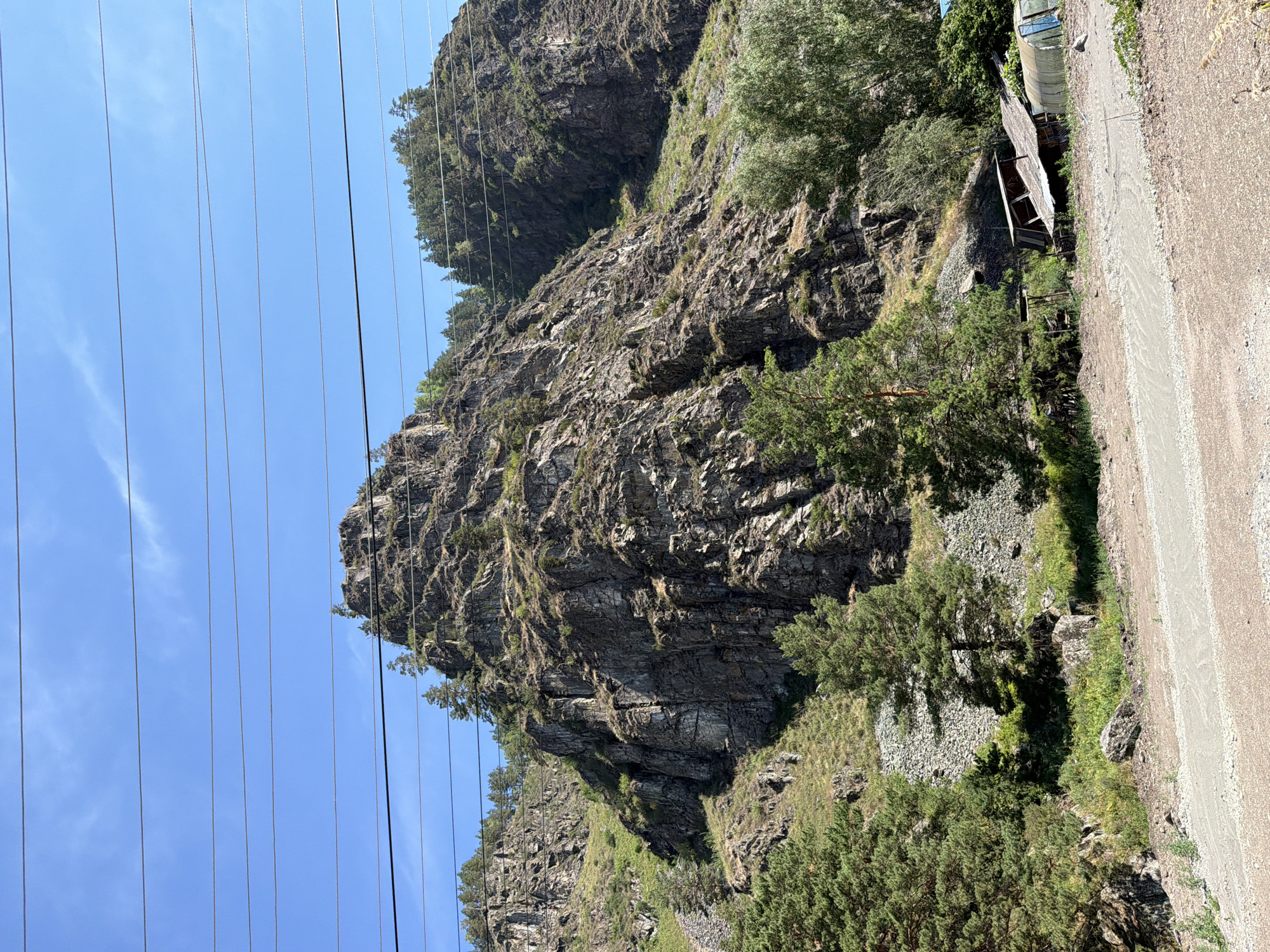 The image depicts a rugged, rocky mountainside in Ust'-Muny, Russia, under a clear blue sky. Several power lines stretch horizontally across the frame, partially obscuring the view of the mountain. The mountain itself is steep and weathered, with sparse vegetation clinging to its slopes, including several evergreen trees. At the base of the mountain, there is evidence of human activity: a dirt road or path winds through the foreground, and several simple structures, including a dilapidated building with a corrugated roof and a greenhouse, are visible. The overall impression is of a remote, natural landscape with some signs of habitation.