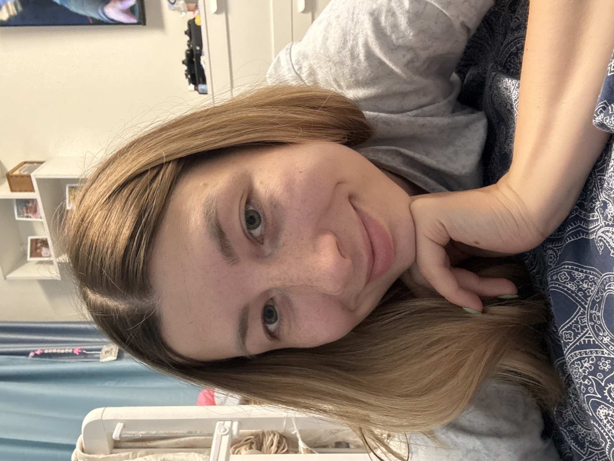 A woman with light brown hair, freckles, and blue-grey eyes lies on a patterned blue blanket, wearing a light gray shirt. Her head is resting on her hand. Behind her is a blue curtain, a white crib, and a white shelf with photos and a small basket. A TV screen hangs on the wall in the background. There is no evidence to suggest that the location is Moscow, Russia.