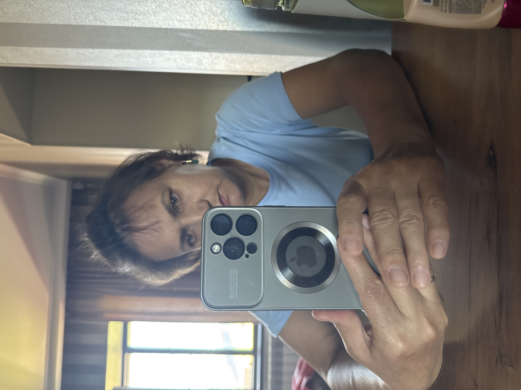 The image shows a woman holding a smartphone, taking a selfie in a dimly lit room with striped curtains and a window in the background. The woman is wearing a light blue t-shirt and has her hair tied back. She is looking at the phone's screen, which reflects her face. The phone case has a circular design with an apple logo. There is a wooden table in the foreground. The specific location in Soloneshnoye, Russia, cannot be determined from the image.