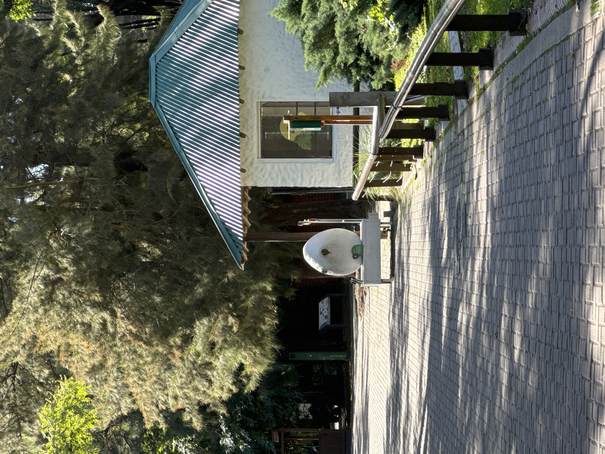 The image depicts a scene in Buenos Aires, Argentina. The foreground showcases a gray brick pathway, diagonally oriented to the viewer. To the left of the pathway is a building with a white exterior and a green, slatted roof. A window is visible, with some kind of design in the reflection. There are railings along the pathway. Beyond the building, there are dense green trees and vegetation, creating a shaded, natural environment. A sign is visible in this shaded area.