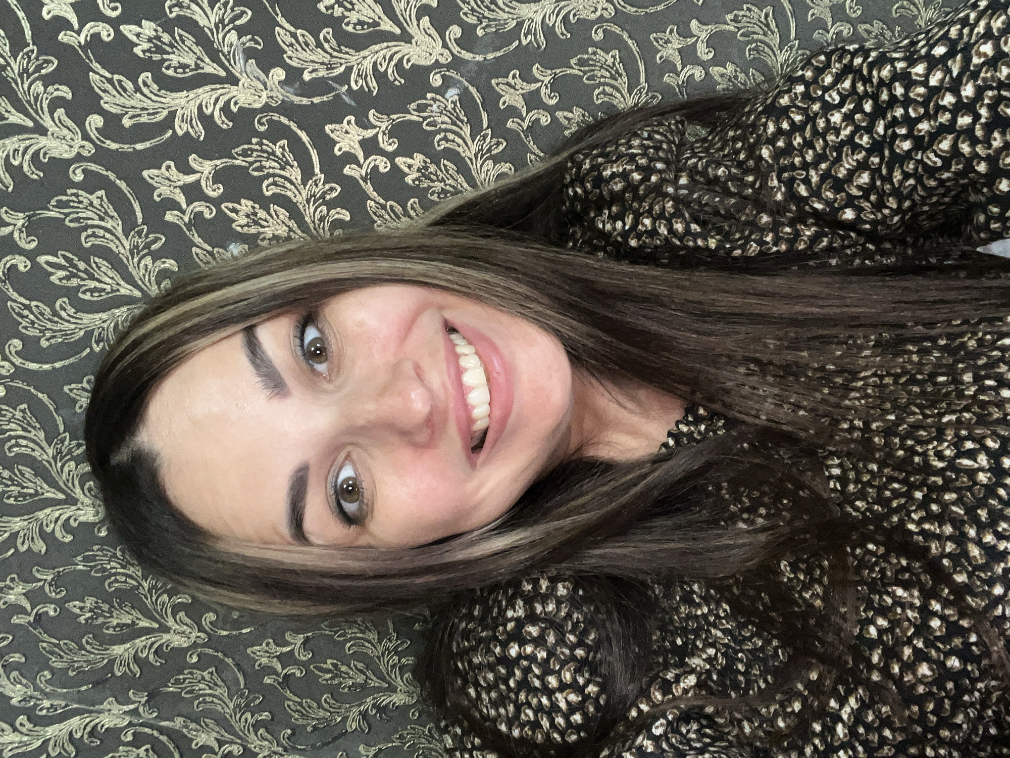 A medium close-up selfie shows a woman with long, dark brown hair with blonde highlights, a wide smile, and brown eyes. She is wearing a black and gold animal-print top. The background is a dark gray damask-patterned wallpaper. The lighting is soft and even. The woman is looking directly at the camera.