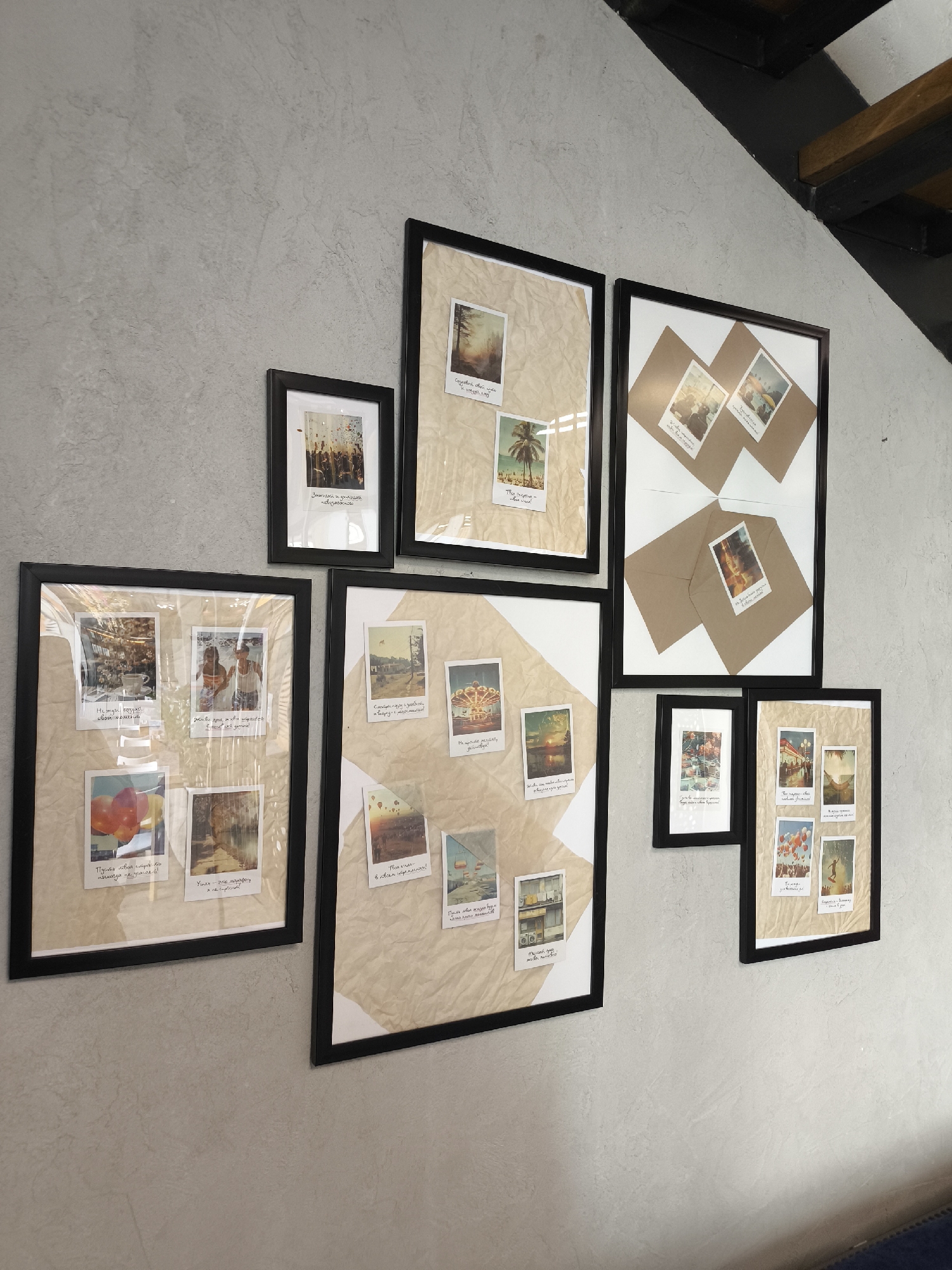 A collection of seven framed photographs is mounted on a light gray wall. The frames are all black and appear to be of varying sizes. The photographs within the frames are arranged on pieces of textured, off-white or light brown paper, reminiscent of parchment. Some frames have a single sheet of this paper as a background, while others have it layered with white paper in geometric patterns.
The photographs themselves seem to be of various subjects, including landscapes, cityscapes, and personal moments. Some are accompanied by handwritten notes in a cursive script.
The wall has a textured finish. In the upper right corner, there is a glimpse of wooden structural elements, possibly stairs or beams.