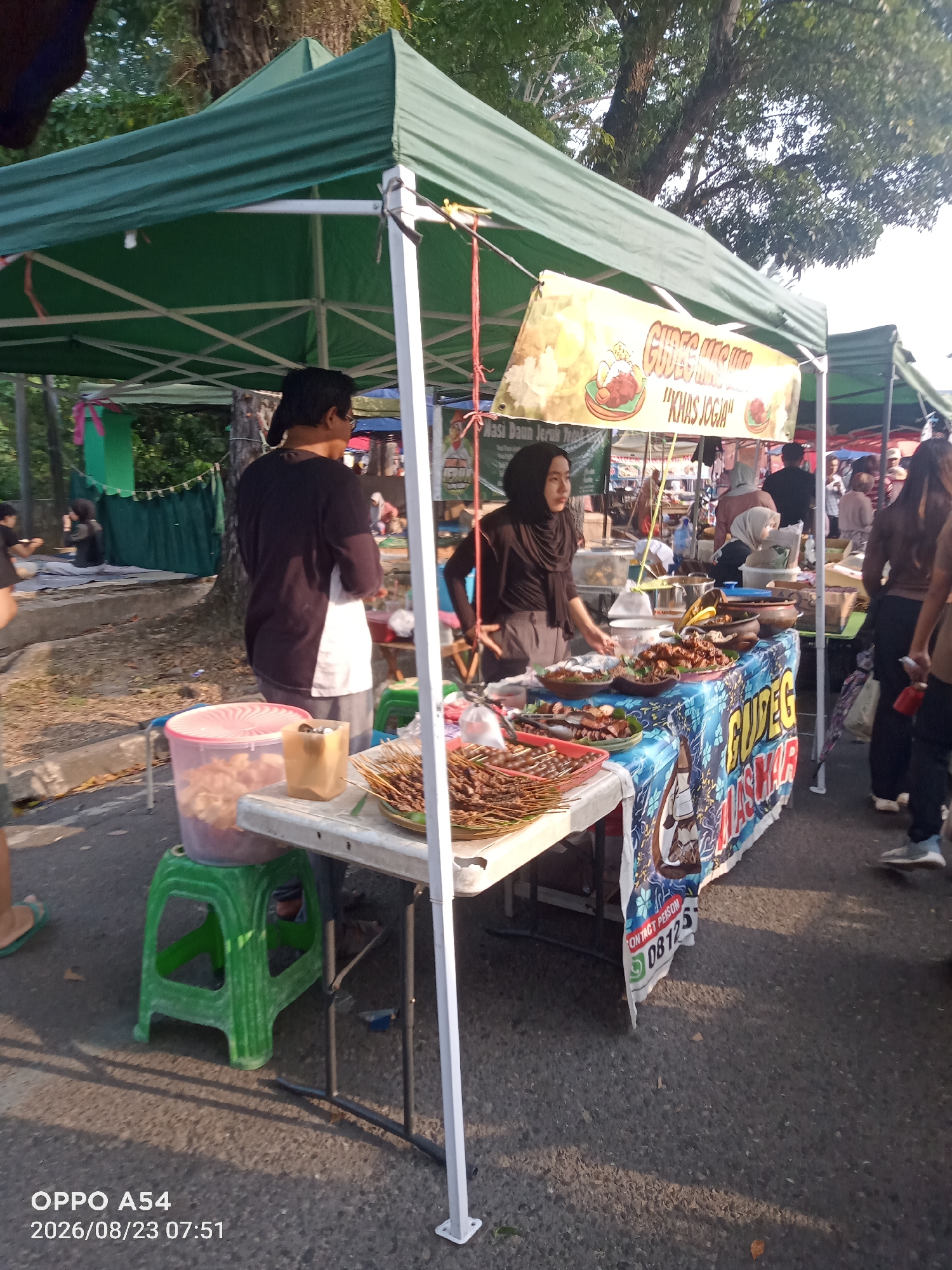 An outdoor food stall selling Gudeg, a traditional Javanese dish, is set up under a green canopy. Two individuals, one male wearing a black long-sleeved shirt and grey pants, and one female wearing a black hijab and grey pants, are attending the stall. The female appears to be serving food. Various dishes, including skewers of meat and what appears to be Gudeg, are displayed on the counter, which is covered with a blue and white patterned cloth. A banner above the stall reads "Gudeg "Khas Jogja"". In the background, other stalls and people are visible, suggesting a market or event setting. The ground is paved with asphalt, and trees can be seen in the background, indicating an outdoor environment. The image is dated 2026/08/23 at 07:51, with the phone model "OPPO A54" also indicated.