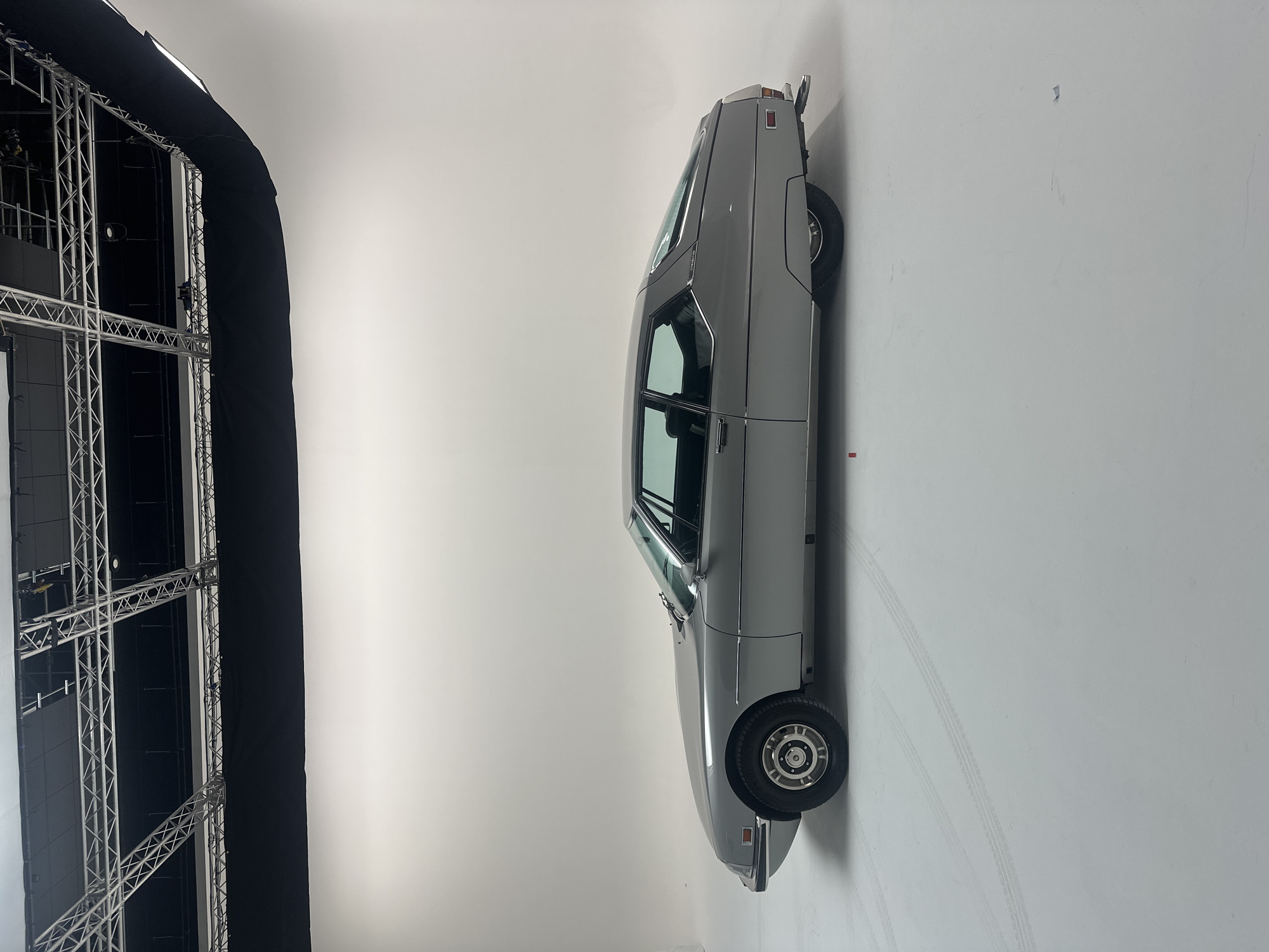 A silver-grey vintage car, identified as a Citroën SM, is the central subject of the image, positioned within a professional photography studio. The car is a two-door coupe, shown from a side profile with its front facing the left side of the frame. It exhibits classic lines, a distinctive long hood, chrome trim around the windows and bumpers, and wheels with silver hubcaps. Faint "SM" badging is visible on the rear window trim. Tire marks are evident on the white floor behind the vehicle, suggesting it was driven into position for the shoot.
The setting is an indoor studio with a large, seamless white cyclorama background that curves upwards from the floor, creating an infinity wall effect. To the left of the car, an extensive metal truss structure, partially covered with black fabric, stands prominently. This structure is indicative of professional staging or lighting equipment, with some studio lights or softboxes visible along its upper edge. The studio is brightly and evenly lit, typical of a controlled environment for product photography or display. No people are visible in the scene. The car is stationary, suggesting an event or activity related to a photoshoot or exhibition. The context provided specifies the location as London, United Kingdom.