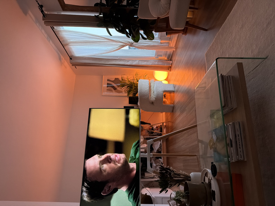 This image displays a modern residential living room interior located in Paris, France. The scene is illuminated by warm, orange-toned ambient light reflecting on light-colored walls and a wooden floor. A large, flat-screen television, positioned centrally on a light-colored tripod stand, shows a male subject with short dark hair and a green shirt, looking towards the left with an open-mouthed, engaged expression. A blurred yellow lamp is visible on the television screen's background. In the foreground, a rectangular coffee table with a clear glass top and a wooden lower shelf holds stacks of magazines, a red box, a potted plant, and dried botanicals in a white vase. To the right of the television, a white, curved, boucle-textured chair with a small cushion rests on the wooden floor. Adjacent to it, near a large window covered by sheer, tied-back curtains, a spherical orange glowing lamp is situated. A light-colored armchair with a visible wooden frame is partially present on the far right. Multiple large potted plants are positioned throughout the room, and a framed abstract art piece hangs on the wall. A rectangular mirror with a wooden frame is partially visible on the far right wall. The specific street or arrondissement within Paris is not visually identifiable.