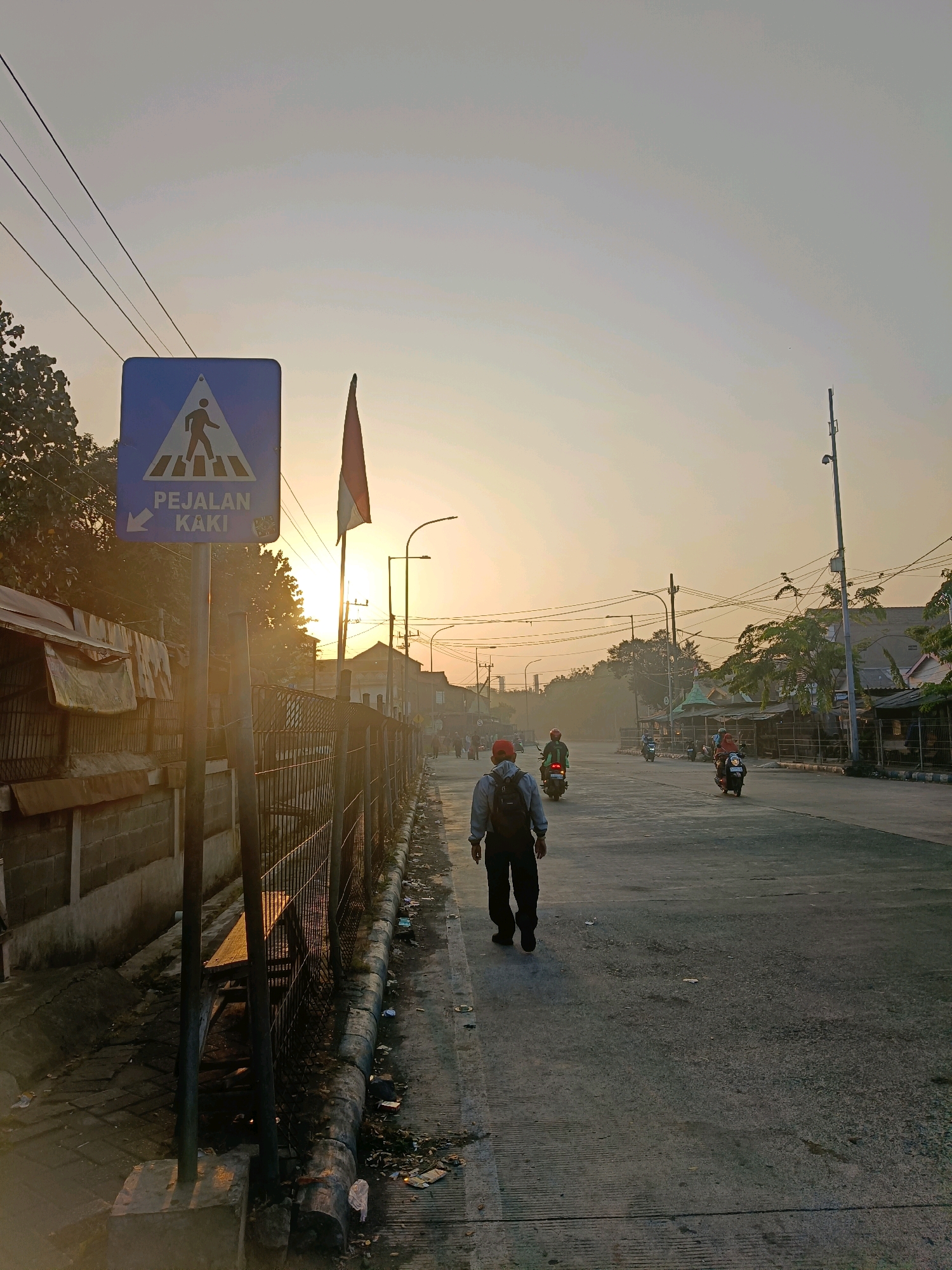 The image depicts a street scene in Kedungrejo, Indonesia, during sunrise. A blue pedestrian crossing sign with the words "PEJALAN KAKI" (Indonesian for pedestrian) and an arrow pointing left is prominently displayed in the foreground. Behind the sign, a tall flagpole with the Indonesian flag is partially visible, silhouetted against the rising sun. The sun's rays create a bright glow, illuminating the scene and casting long shadows.
The street is paved and appears to be a main thoroughfare. Several people on motorcycles are traveling along the road. In the mid-ground, a person wearing a red cap and a backpack is walking away from the camera, towards the intersection. A wire fence runs along the left side of the road, separating it from a sidewalk area. This area appears somewhat unkempt, with debris on the ground. Buildings and trees line the sides of the street. The sky is hazy and light orange, characteristic of a sunrise.