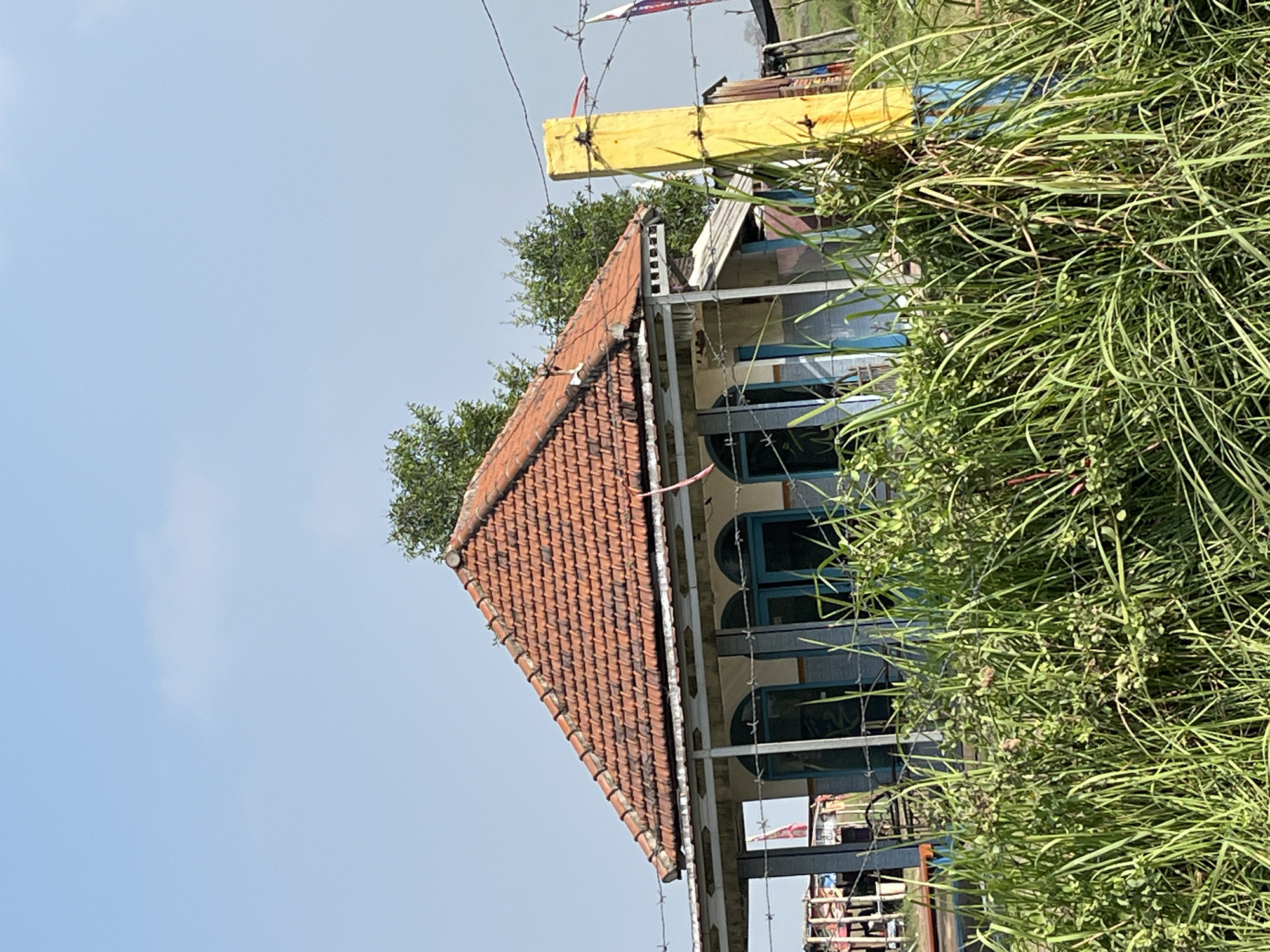 The image captures a scene in Gempolsari, Indonesia, featuring a small, weathered building with a red-tiled roof and blue-trimmed windows. The building appears to be situated in a rural or semi-rural environment, with tall green grass and vegetation in the foreground. A yellow post, possibly part of a fence, stands prominently on the right side of the frame, with barbed wire strung across it and around the scene. The sky is clear and blue, indicating daytime. The building's architecture suggests a utilitarian purpose, possibly a dwelling or a small community structure. The overall impression is one of simplicity and rural life, with elements of weather-worn structures and natural growth. There are no people visible in the image.