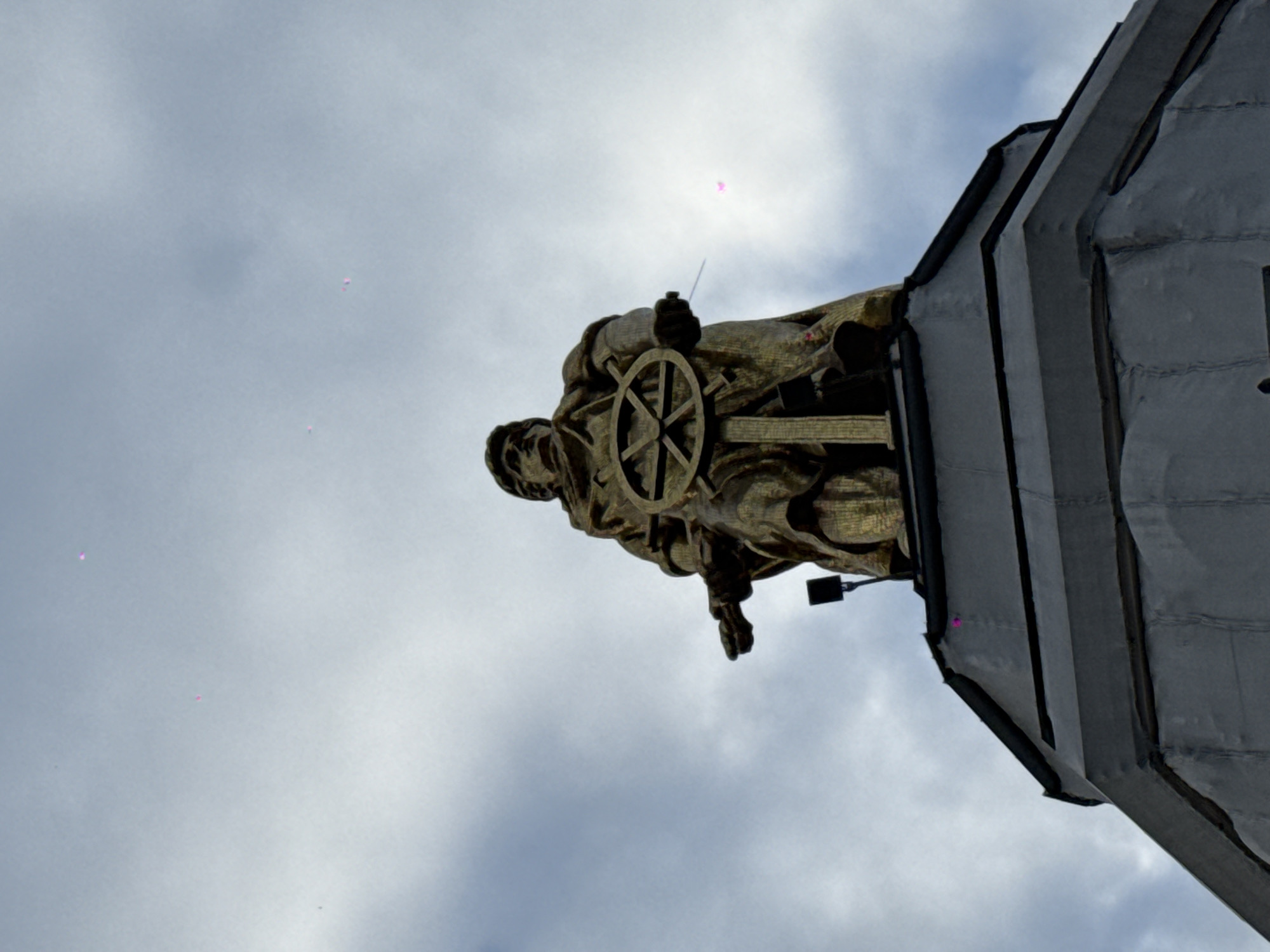 A low-angle shot depicts a golden statue of a man holding a wheel, possibly a saint, mounted atop a gray, tiered stone structure against a cloudy sky. The statue is weathered with visible details in the clothing and facial features. The sky has scattered pink highlights. The location is likely a monument in Moscow, Russia.