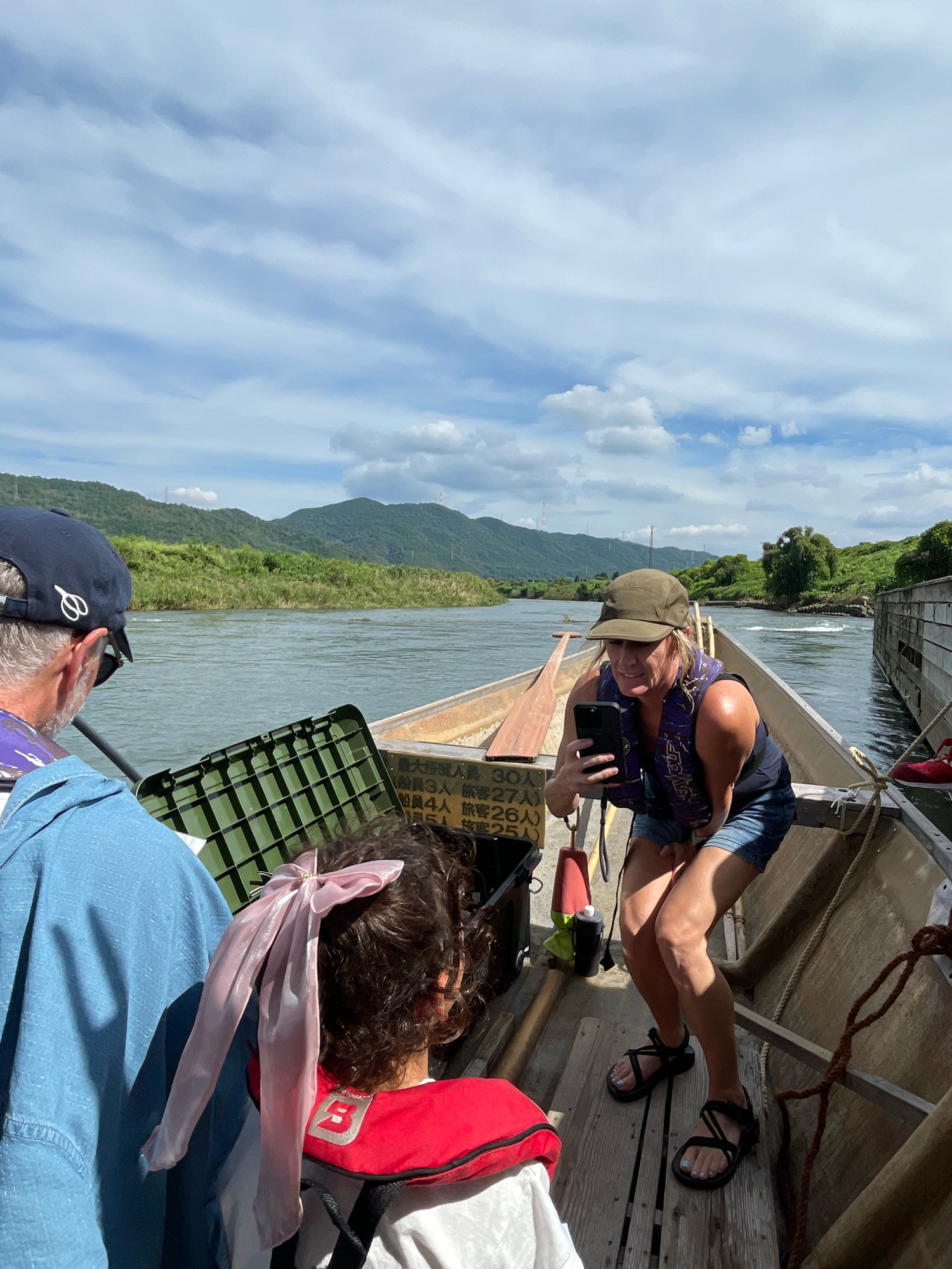 In Kameoka, Japan, on a sunny day with scattered clouds, people are on a wooden boat traveling down a river. A woman in a green hat and life vest is taking a selfie with her phone. A man with a blue hat is looking out at the river, and a child with a pink bow in her hair is wearing a red life vest. The river is surrounded by lush green mountains. The boat is equipped with oars and life vests, suggesting a guided tour or activity.