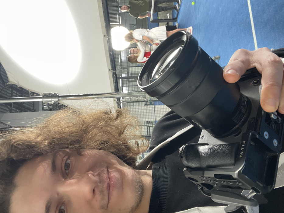 The image captures an indoor padel court scene within a facility located in Yekaterinburg, Russia. In the immediate foreground, a male individual with medium-length wavy brown hair and light stubble, wearing a black long-sleeve top, holds a black Sony mirrorless camera, specifically a model fitted with an E PZ 18-110mm F4 G OSS lens and mounted within a SmallRig TA-T13 camera cage. He holds the camera in a selfie-like manner, looking towards the lens with a neutral expression, a camera strap visible over his left shoulder.
The background reveals a bright blue padel court with white boundary lines and a black net. Three individuals are positioned to the left of the net: two women, one in a white dress with red accents and the other in a white dress, are engaged in an interaction, possibly posing or having clothing adjusted. A third male individual, wearing glasses, is partially visible between the two women, facing the woman in white. To the right of the net, a fourth male individual, dressed in a dark green t-shirt and dark pants, stands with his hands in his pockets, observing the scene.
The area is illuminated by professional studio lighting, including a large, curved white overhead diffuser and a smaller, spherical softbox positioned on a stand to the left of the net. Metal grid walls, typical of a padel court, and overhead structural elements are visible in the background. A single yellow padel ball rests on the blue court surface near the net.