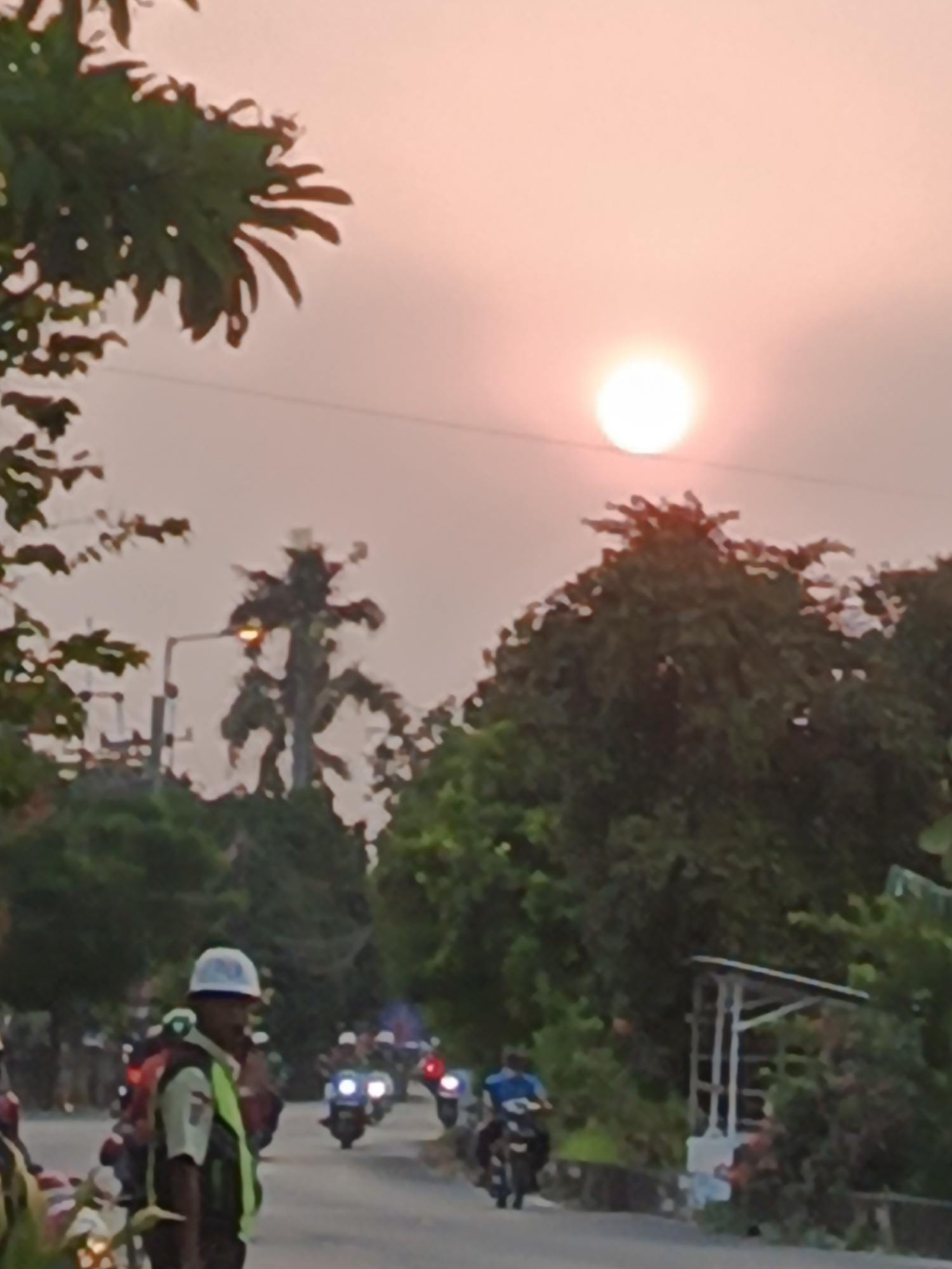 The image depicts a street scene at dusk in Wedi, Indonesia, with the sun partially visible in the hazy sky. In the foreground, a person wearing a white helmet and a reflective vest is standing by the side of the road. Behind this individual, a procession of motorcycles is proceeding along the paved road, with several vehicles displaying illuminated headlights and taillights. The road is flanked by lush green trees and vegetation. A street light is visible on the left side of the image. The overall atmosphere suggests a departure or arrival activity involving the motorcycles and the individual in the foreground.
