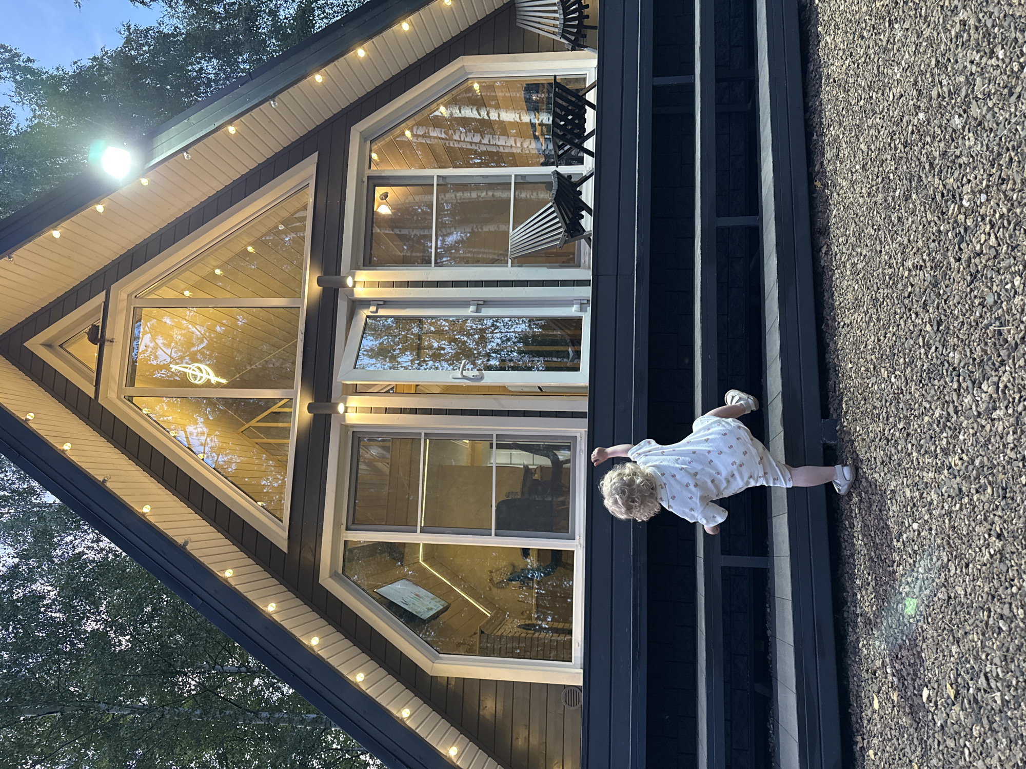 In Prokudino, Russia, a small child with curly blonde hair, wearing a light-colored outfit with a floral pattern and white sandals, is climbing up a dark set of outdoor stairs leading to a modern, triangular-shaped house. The house has a dark exterior with white trim and is illuminated by string lights along the roofline and a bright spotlight above the large, angular windows. Through the windows, interior lights are visible, reflecting the surrounding trees. Two dark outdoor chairs and a small table are on the porch in front of the house. The ground in front of the stairs is covered in gravel.