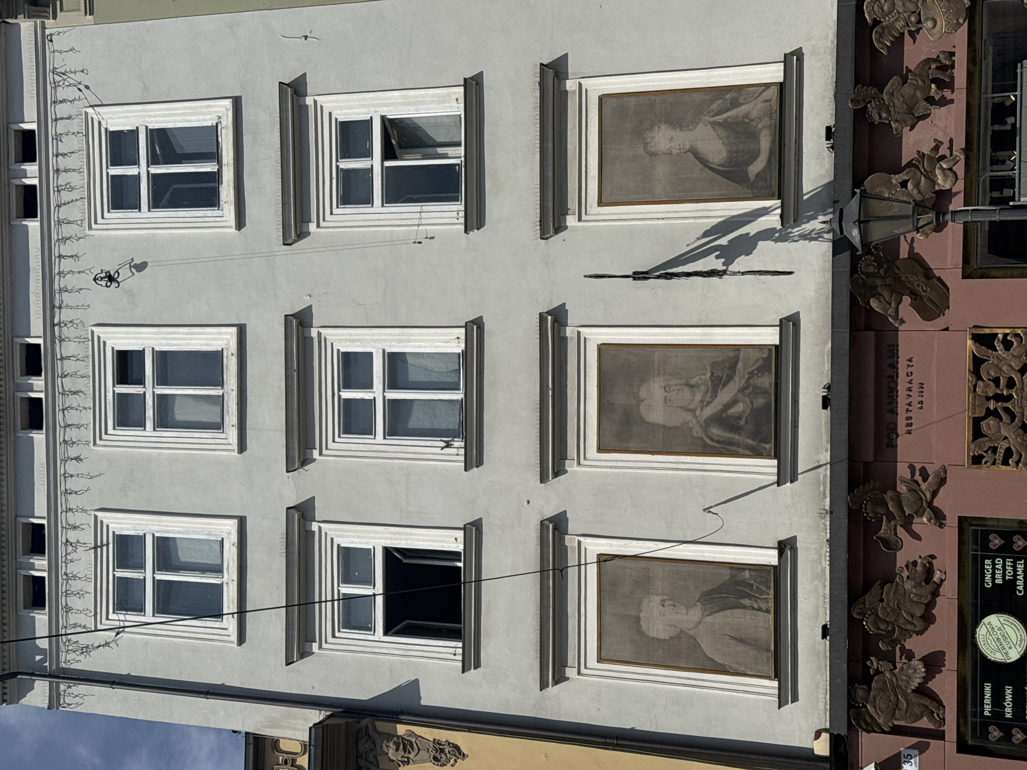 The image shows a close-up view of the facade of a building located in Krakow, Poland. The building is light gray and features a grid of nine windows, each with white frames. The windows are arranged in three rows of three. The top row of windows appears to be open, revealing darkness within. The middle row of windows are closed, with one on the left side appearing to be slightly ajar. The bottom row of windows are not actual windows but framed portraits. The leftmost portrait depicts a man with a powdered wig and formal attire. The middle portrait shows a person in similar attire, and the rightmost portrait displays a woman in a formal gown and hairstyle. Below these portraits, a decorative frieze runs across the building, adorned with relief sculptures of cherubic figures and musical instruments. A street lamp with a black pole and a lantern is positioned in the lower right section of the image, casting a shadow on the building. Signs for "PIERNIKI" and "GINGER BREAD TOFFI CARAMEL" are visible at the bottom of the image, indicating a food establishment. The sky is a clear blue, suggesting it is daytime. The overall architectural style suggests a historic building.