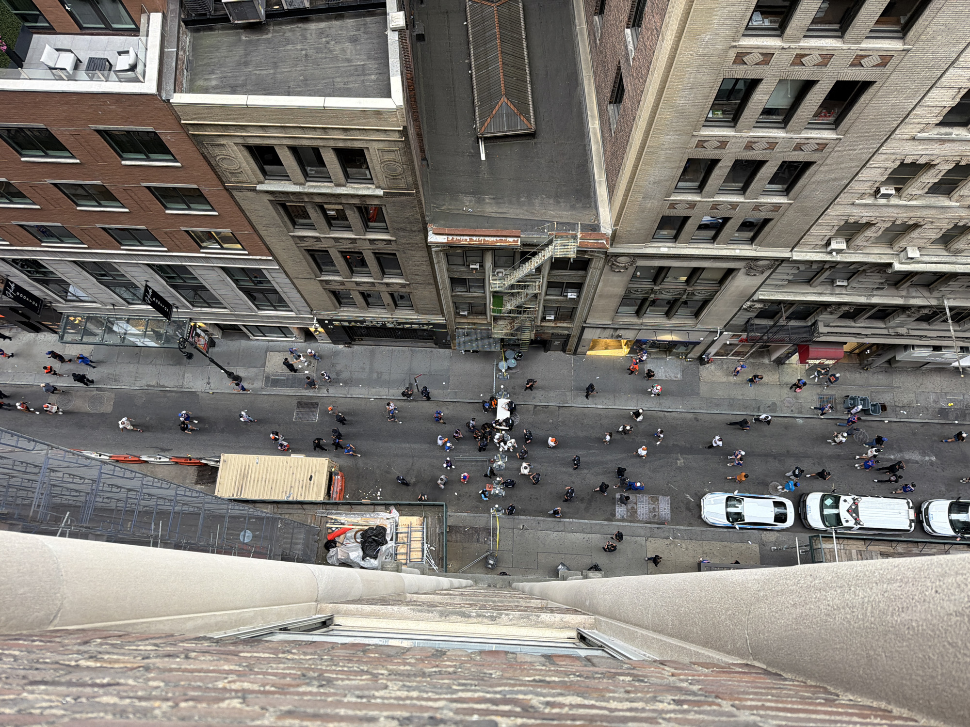 This is a high-angle, top-down shot of a street scene in New York City, United States of America. The perspective is from an elevated position, looking down on a narrow street bordered by multi-story buildings. The street is paved with asphalt and marked with painted lines.
There are several vehicles on the street, including two white vans, one white sedan with a blue stripe, and a white SUV. The vehicles are parked and appear to be operational.
A significant number of people are walking in the street and on the sidewalks. Many of them are walking in groups, while others are alone. Some individuals are wearing casual clothing, while others appear to be in work attire. There are also some people gathered in small clusters.
The buildings lining the street are of various architectural styles and materials. One building on the left has a brick facade and a stone decorative element. Other buildings have a mixture of brick and concrete, with numerous windows. Some buildings have external metal staircases. There are also signs and awnings visible on some of the buildings.
Scaffolding and construction materials are present on the side of one of the buildings, suggesting ongoing renovation or construction work. A large shipping container is also visible on the street.
The environment appears to be a busy urban setting, with signs of commercial activity and pedestrian traffic. The lighting suggests it is daytime. The overall scene depicts a lively street with a mix of vehicular and pedestrian activity, set against the backdrop of dense urban architecture.