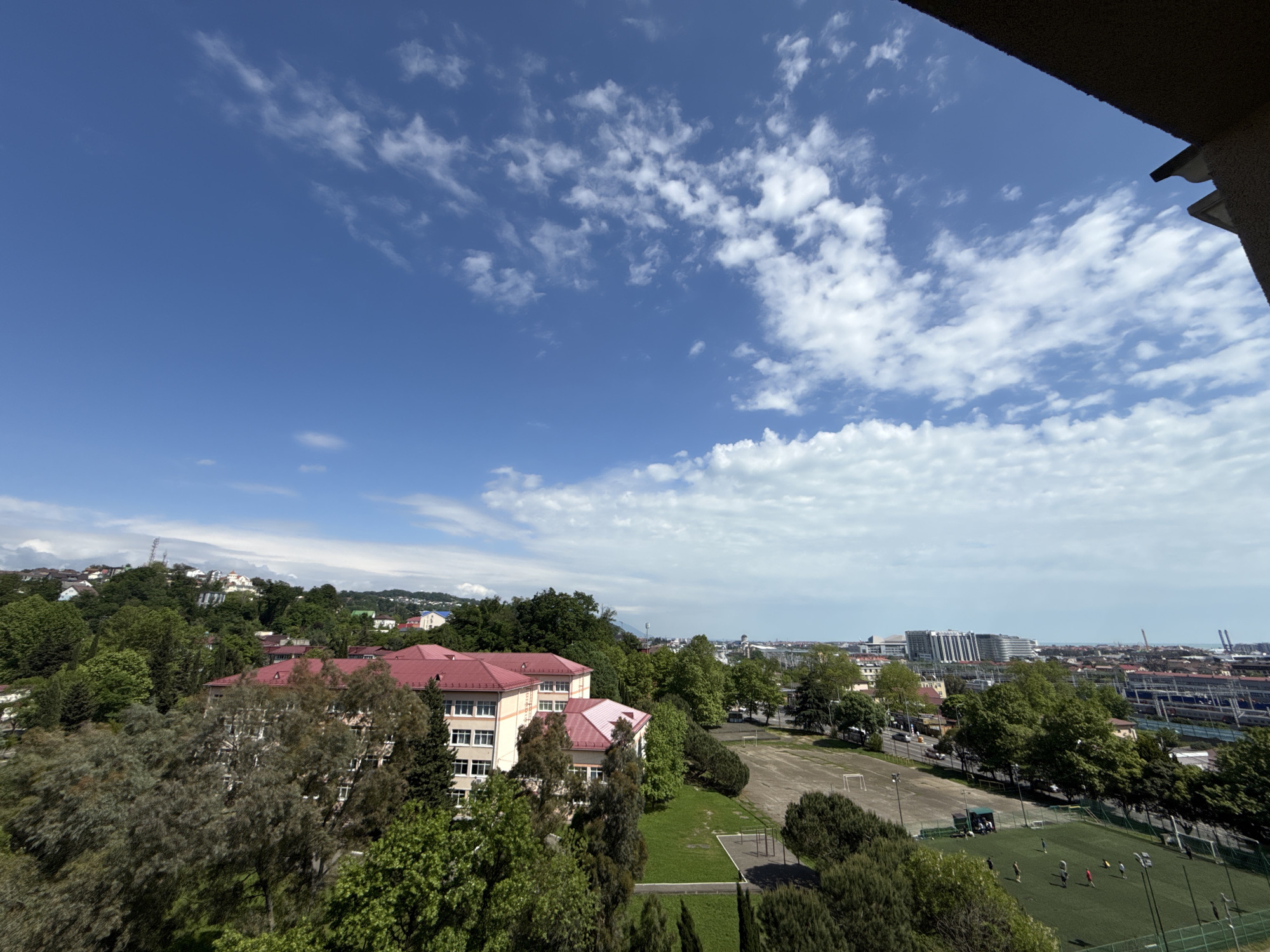 The image depicts a view overlooking a part of Sochi, Russia. In the foreground, a large building with a red roof is partially visible, surrounded by dense greenery. Further away, a sports field is seen, with people engaging in some form of activity, possibly a soccer game. Beyond the sports field, the cityscape of Sochi stretches into the distance, punctuated by various buildings and structures. A clear blue sky with scattered clouds is overhead. The composition suggests the image was taken from a high vantage point, possibly from a building or an elevated location.