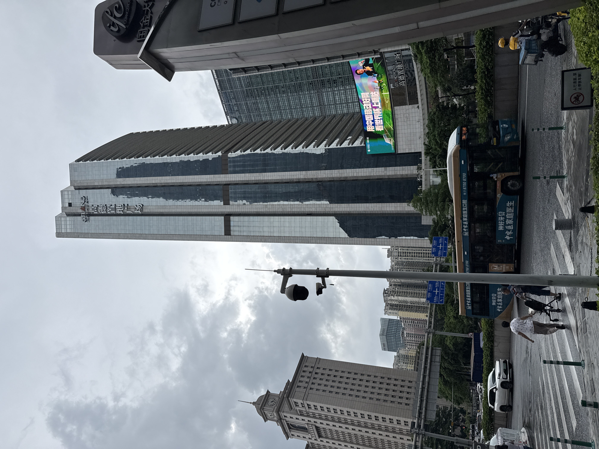 In Tianhe District, China, a cloudy, overcast day is depicted. The scene is dominated by modern skyscrapers and a prominent billboard displaying an advertisement. A bus is stopped at a crosswalk, and pedestrians are crossing the street. A white SUV is also visible, and a motorcycle with a delivery box is parked nearby. A CCTV camera is mounted on a pole. The overall impression is one of urban bustle under a muted sky.