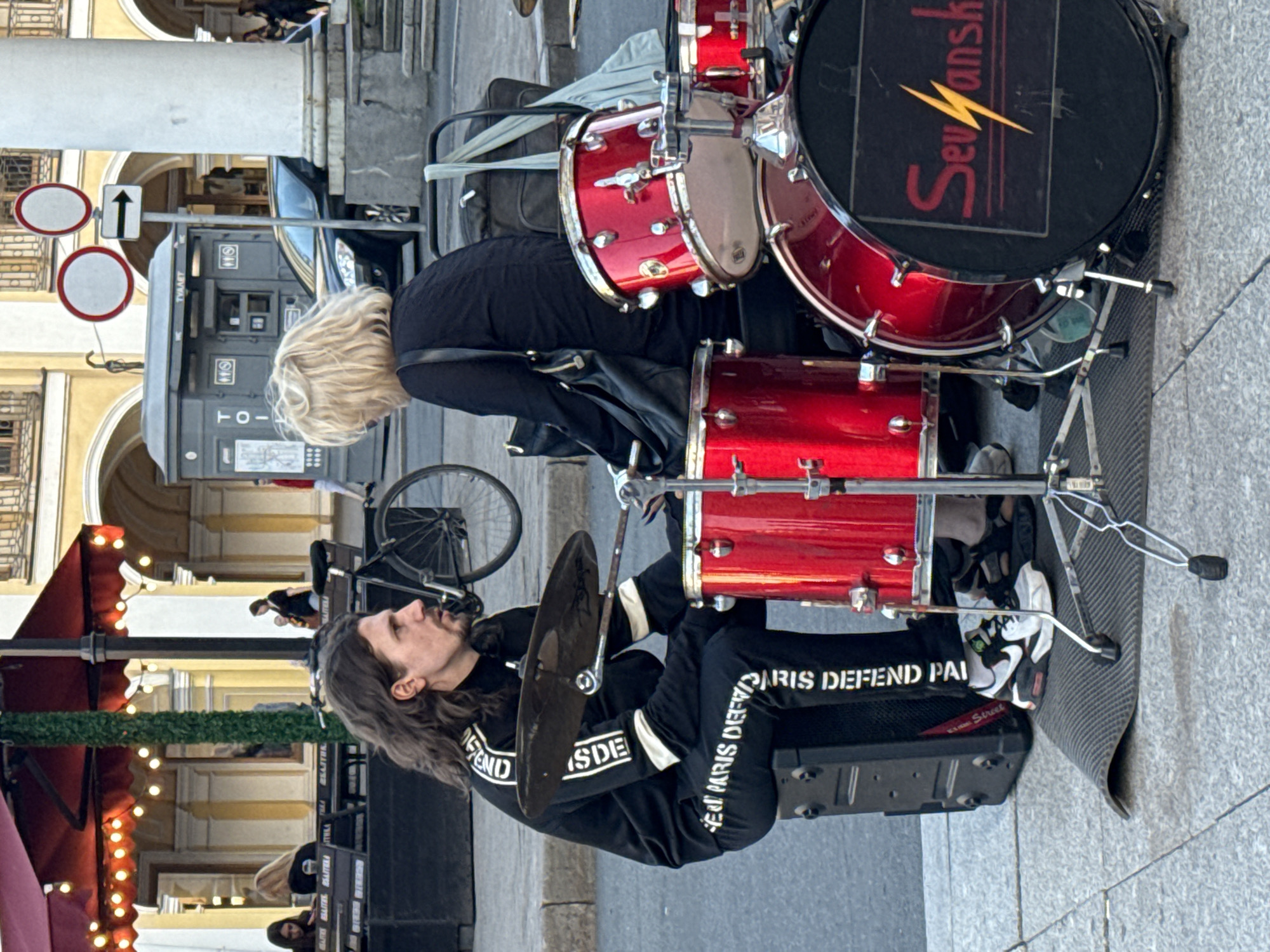 This image depicts a street scene in Saint Petersburg, Russia, possibly on Nevsky Prospekt. In the foreground, a man with long brown hair and a goatee, wearing a black tracksuit with white lettering that reads "PARIS DEFEND PARIS DEFEND," is seated behind a red drum kit. He is in the process of playing the drums, with a cymbal positioned above the snare drum. To his right, a woman with blonde hair, wearing a black top and carrying a black handbag, is leaning towards him, seemingly engaged in conversation. Behind the musicians, a black car is parked, and further in the background, a public toilet facility and several people can be seen. The overall environment suggests an outdoor urban setting with architectural elements and street furniture.