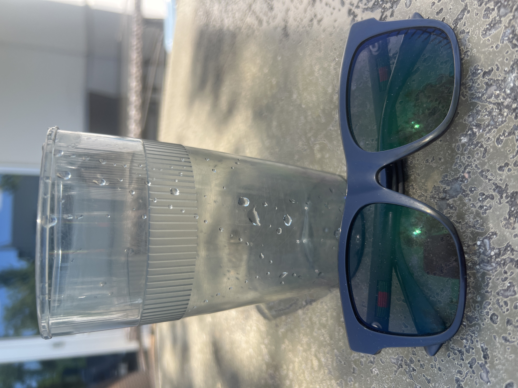 A pair of dark blue sunglasses rests on a textured, light-colored surface in the foreground. Behind the sunglasses, a clear plastic cup filled with a light-colored liquid is positioned slightly to the left. The exterior of the cup is covered in condensation. The background is blurred, showing what appears to be a building with white siding and reflections of trees and sky in its windows. The overall lighting suggests an outdoor setting on a sunny day.