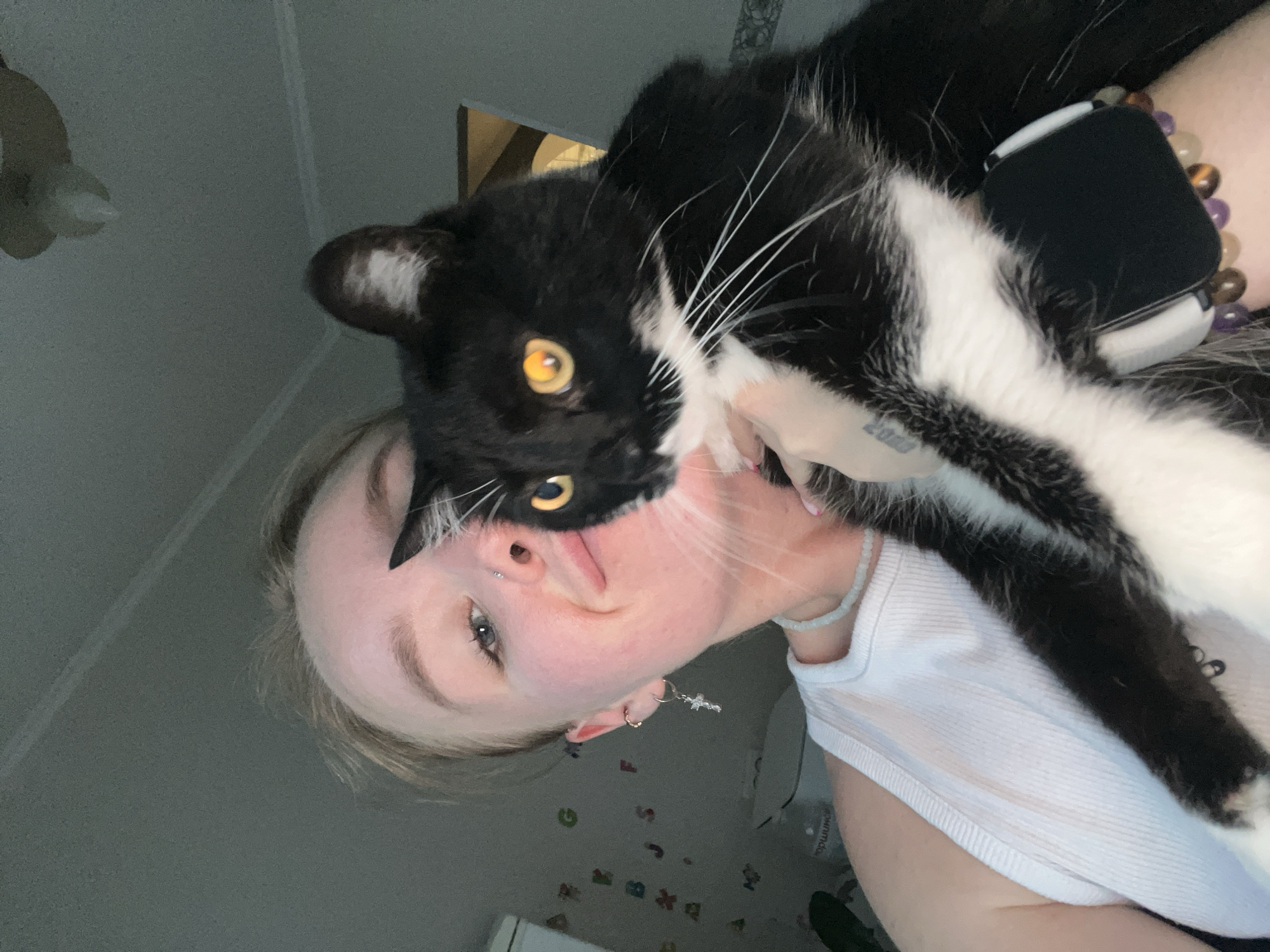 A close-up, overhead shot features a person and a black and white cat. The person, facing upwards, has blonde hair and is wearing a white tank top. Their left cheek is pressed against the cat's fluffy white fur. The cat, with striking yellow eyes, is positioned above the person's face, its head tilted downwards. The cat's black fur is interspersed with white patches on its chest and paws.
The background appears to be an indoor setting, possibly a bedroom, with a grey wall and some objects visible on it, including what looks like stickers of letters and numbers. A light fixture can be seen in the upper left corner. The lighting suggests it might be nighttime or a dimly lit room. The overall tone of the image is intimate and affectionate, conveying a sense of closeness between the person and their pet. The person has several piercings, including one on their nose and another on their ear. They are also wearing a delicate necklace and earrings. A beaded bracelet with a smartwatch is visible on the person's wrist. No weather or specific time of day cues are evident beyond the indoor lighting. No visible text is present in the image.