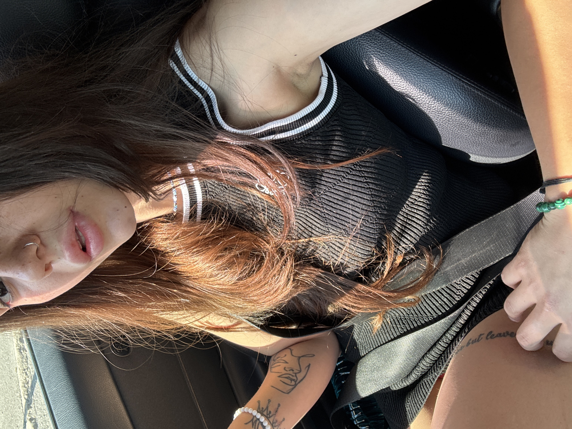 A young woman with long, brown hair and a nose ring sits in the passenger seat of a car. She is wearing a black tank top with white trim. Her left arm, which is visible, has tattoos and a beaded bracelet. A seatbelt is fastened across her chest. The interior of the car appears to be black leather. The lighting suggests it is daytime, with sunlight casting shadows. The background outside the car window is blurred, but appears to be a sandy or dusty surface. There is no information in the image to suggest a location in Sofia, Bulgaria.