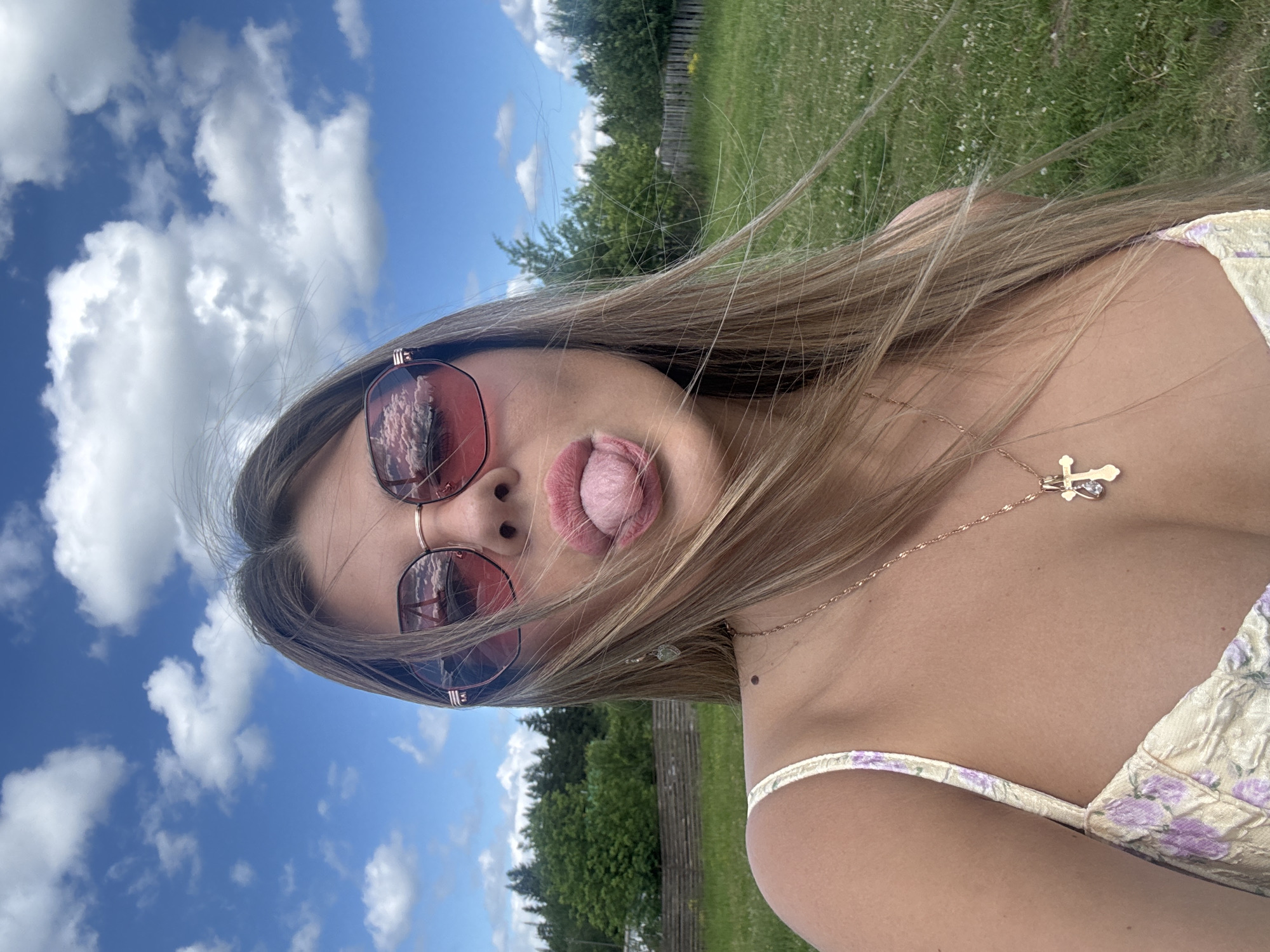 The image shows a young woman taking a selfie outdoors. She is wearing pink-tinted sunglasses and a yellow floral top. Her hair is long and blonde, and it is blowing in the wind. She is sticking her tongue out, and her lips are slightly parted. In the background, there is a blue sky with white clouds, green grass, and trees. The time of day appears to be daytime, and the weather is sunny and clear. The overall mood of the image is playful and casual. The woman is wearing a gold chain with a cross pendant. There are no visible text elements in the image. The location is identified as Pastukhov, Russia.