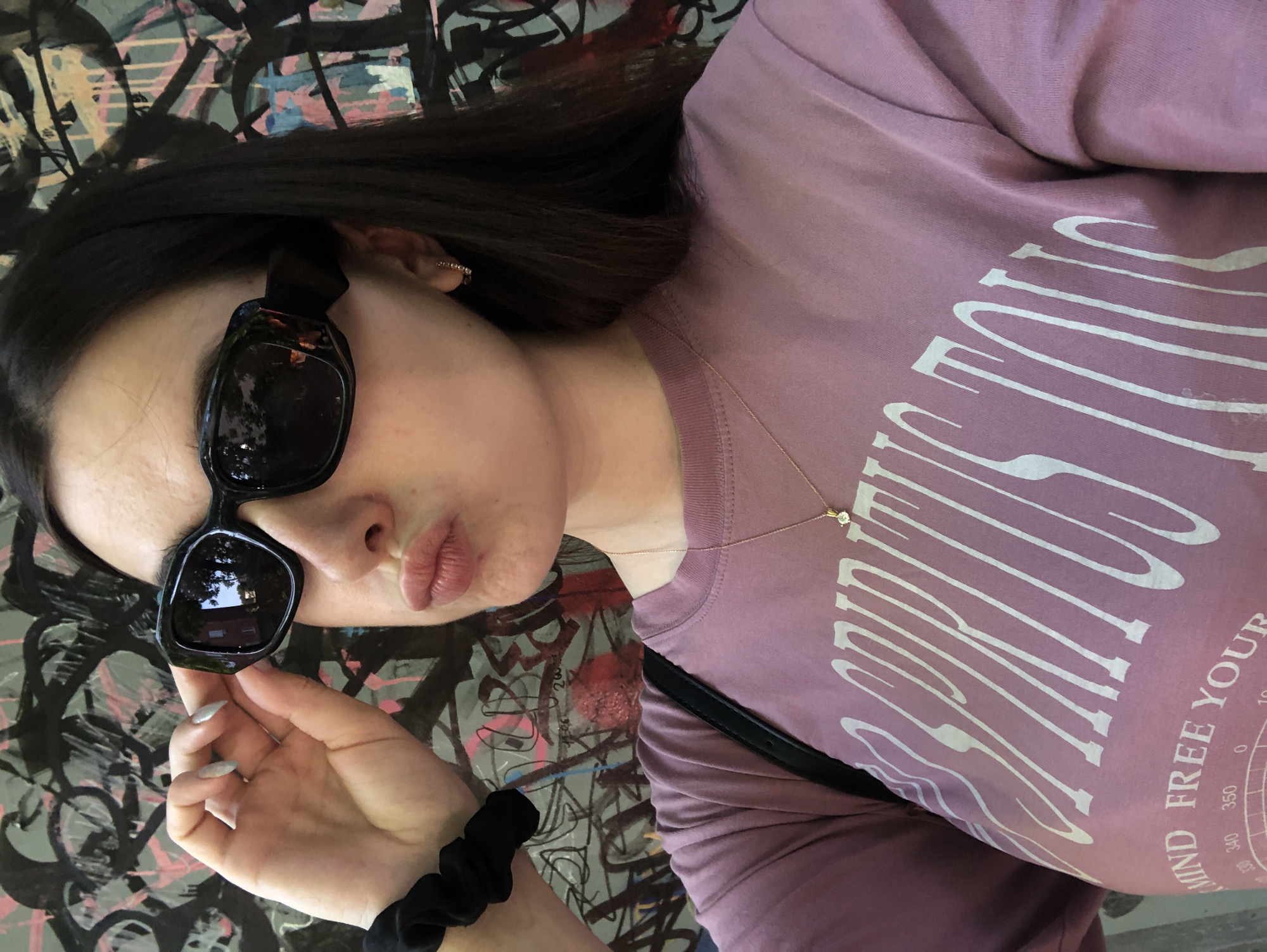 A selfie of a woman is in front of a graffitied wall. The woman is fair-skinned with dark hair and is wearing sunglasses, a lavender t-shirt with white stylized text that reads "MIND FREE YOUR...," and a delicate gold necklace. She is holding her sunglasses with one hand, and a black scrunchie is visible on her wrist. The background consists of graffiti art with a mix of colors including pink, grey, and black.
The location of the city of Chaikovsky, Russia, cannot be determined from the image.