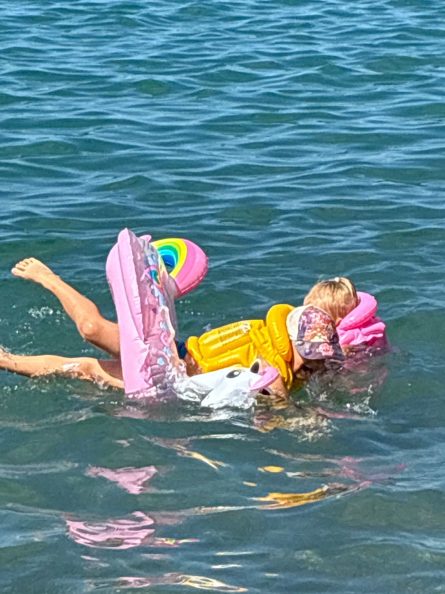 The image depicts a person in the water, wearing a yellow life jacket and resting on a pink inflatable raft. The raft has a rainbow design and appears to be shaped like a unicorn. The person's legs are visible kicking in the water, and they are partially submerged. The water is choppy and a deep blue-green color. The scene is set in the open water, with no land visible. The location is not specified in the image or provided context.