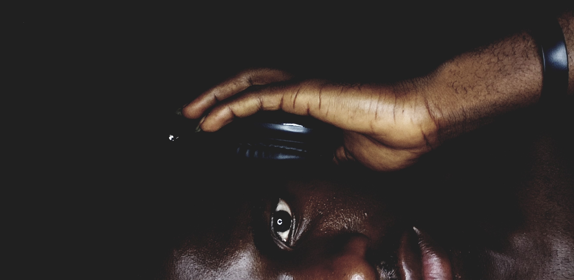A dimly lit, close-up shot shows a dark-skinned person from the chest up. The person's face is angled upwards, with only one eye clearly visible. A hand is positioned above the person's face, resting on a black object. The location is unknown and there is no evidence to suggest that this photograph was taken in Narayi, Nigeria.