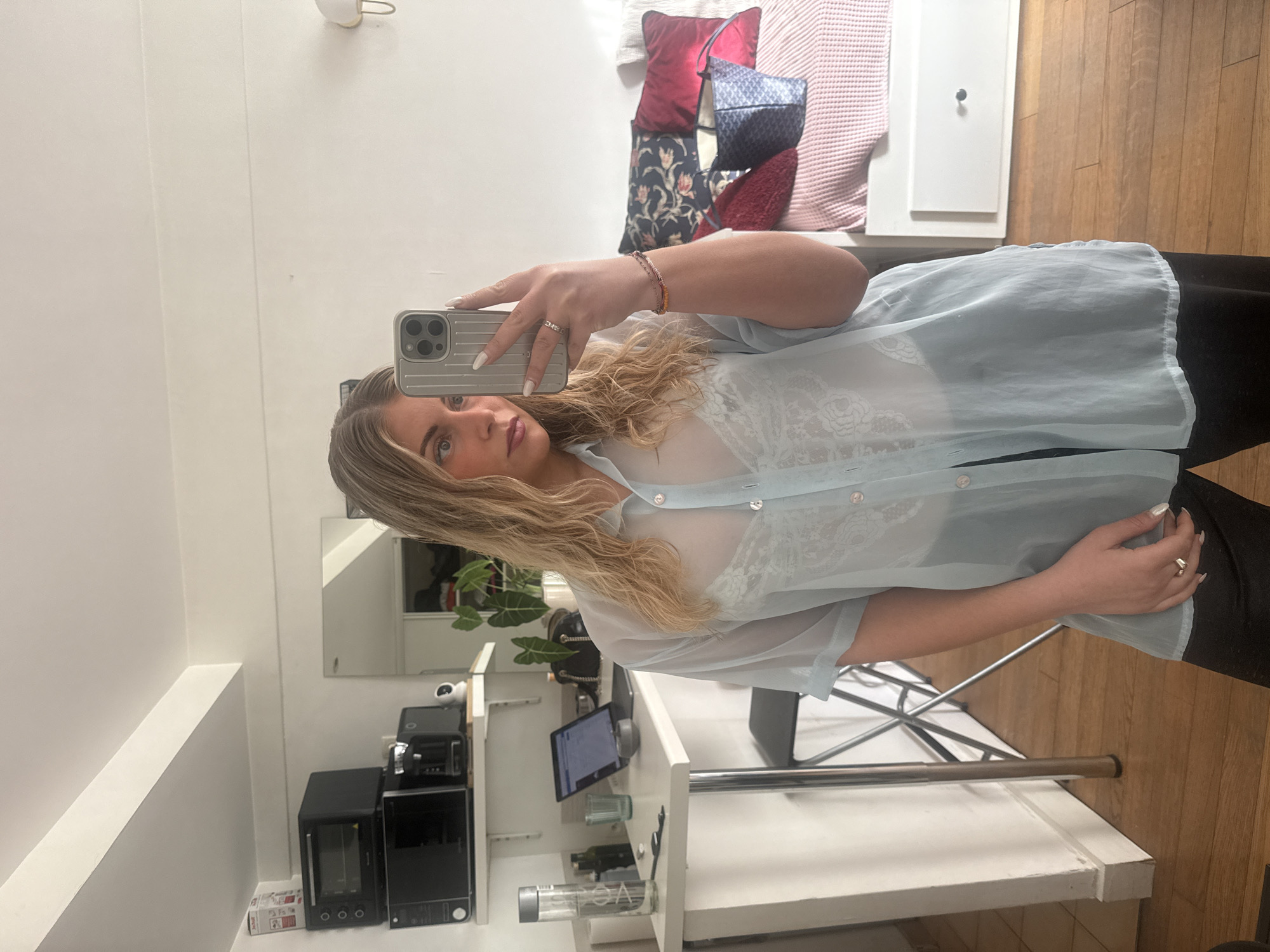 A woman is taking a selfie in a modern apartment with white walls, possibly in Paris, France. She's holding a silver iPhone in her left hand, showcasing her blonde wavy hair and a light blue, sheer button-up shirt with a lace bra underneath. Her right hand is visible near her hip, displaying a ring. She is wearing black pants. Behind her, there's a kitchen area with a microwave, oven, security camera, and an iPad on a white counter. In the background, there's a living area with pillows on a daybed and wooden flooring. The apartment is well-lit.