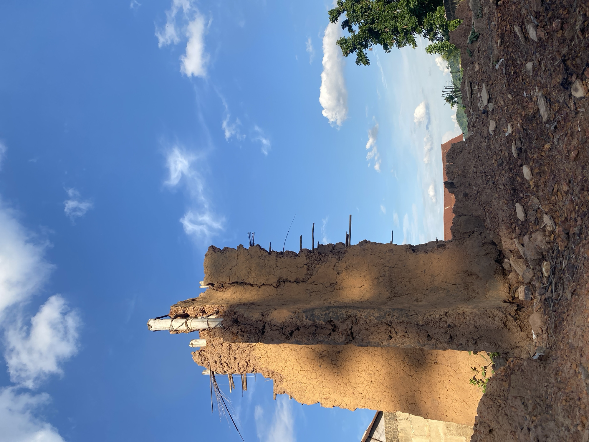 The image shows a deteriorated mud wall structure in Ufu Esioni, Nigeria. The wall is made of packed earth or clay, exhibiting extensive cracking and erosion. Remnants of wooden sticks or posts are visible protruding from the top of the wall, suggesting a former roof or upper structure. The wall's state indicates disrepair or abandonment. In the background, there's a partially obscured building and vegetation. The sky is visible above, with scattered clouds. The image is oriented in an unusual way, possibly rotated.