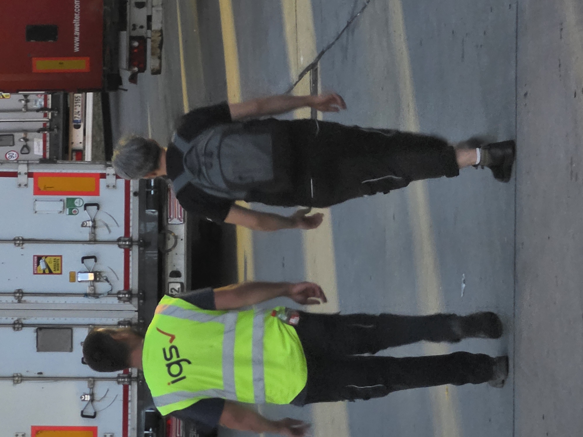 The image depicts two individuals walking away from the camera on a paved surface, possibly in an industrial area or logistics hub. In the background, a large white truck trailer with red and yellow markings is visible, along with part of a red truck trailer. The ground is a flat, grey concrete surface with some faint yellow lines and shadows.
The person on the left is wearing a bright yellow reflective vest with "igs" and a red checkmark logo on the chest. Underneath the vest, they are wearing a dark t-shirt and black pants. A clear plastic water bottle is visible tucked into the waistband of their pants. They have dark hair and appear to be looking to their left.
The person on the right is wearing a black t-shirt and black pants. They are carrying a grey backpack. They have short, grey hair. Their gaze is directed forward.
The overall scene suggests an outdoor environment, possibly a loading dock or a service area associated with transportation or logistics. The presence of reflective vests indicates a work environment where safety attire is required. There is no specific visual information to pinpoint the exact location within Frankfurt, Germany.