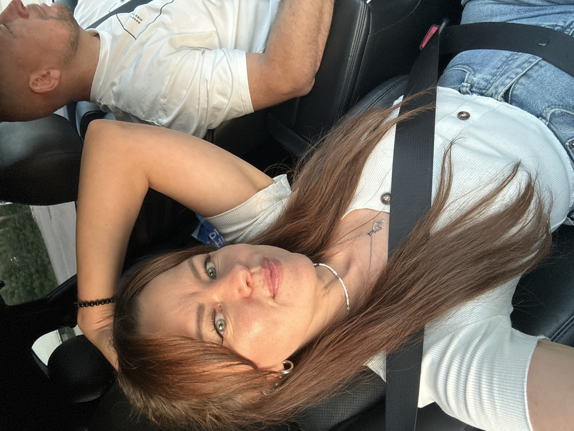 The image shows two people, a man and a woman, sitting in the front seats of a car. The man is in the background, wearing a white t-shirt and is facing away from the camera, his head tilted back. The woman is in the foreground, looking towards the camera with her head tilted. She is wearing a white top and jeans. Both individuals are wearing seatbelts, indicating they are traveling. The scene appears to be set during the daytime, with natural light illuminating the interior of the car. There are no obvious indicators of the weather, but the lighting suggests it is not dark or overcast. The car interior, with its black leather seats and seatbelts, is the primary setting. There is some visible text on the man's t-shirt, but it is blurry and difficult to read. The woman has a necklace on and appears to be relaxed and engaging with the camera. The image seems to capture a casual moment during a car ride.