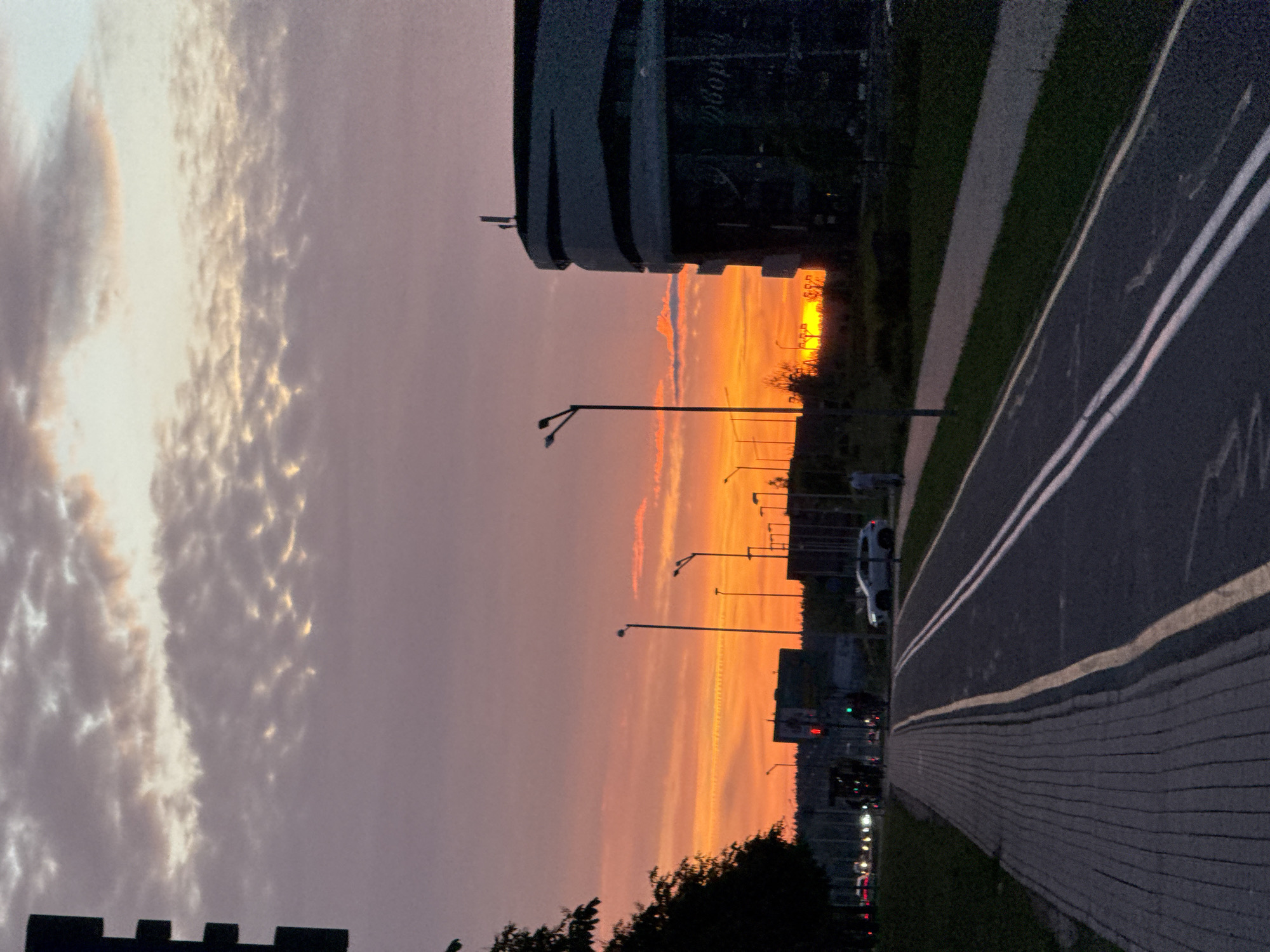 This image captures a wide-angle view of an urban landscape at sunset in Minsk, Belarus, specifically in the vicinity of the Minsk-Arena complex. The sky is dominated by vibrant orange and yellow hues near the horizon where the sun is setting, blending into pink, purple, and light grey clouds higher up.
In the foreground, a dark grey paved pedestrian walkway with a cobblestone-like pattern runs along the left side. Adjacent to it is an asphalt road featuring a dedicated cycling lane marked by a single faded yellow line separating it from the sidewalk and two parallel solid white lines (double solid white) on its other edge. The road surface appears dark.
The midground features various elements:
* On the far left, a dark silhouette of a deciduous tree partially obscures a traffic light.
* Numerous tall, dark street light poles with luminaires are spaced along both sides of the road and a central median.
* Vehicles are visible on the road in the distance, identifiable by blurred headlights and taillights, with at least one white SUV discernible on the right side of the main road.
* Several human figures are present: one person walks on the sidewalk on the far right, another person is further down the road near the left traffic light, and a cyclist is faintly visible near the left edge of the road.
* A green traffic light is active on a pole in the left-center.
* On the right side of the image stands a prominent, large, modern building with a distinctive curved, dark glass facade and a multi-pronged antenna structure on its roof. The building's surface reflects the low light and dark sky. Partially visible Cyrillic text on the facade reads "Дворец" (Palace), indicating a cultural or sports complex. The architecture and visible text are consistent with structures within the Minsk-Arena complex.
The background shows a silhouette of dense trees and distant urban structures against the brightly lit horizon where the sun is setting, casting a brilliant orange glow.