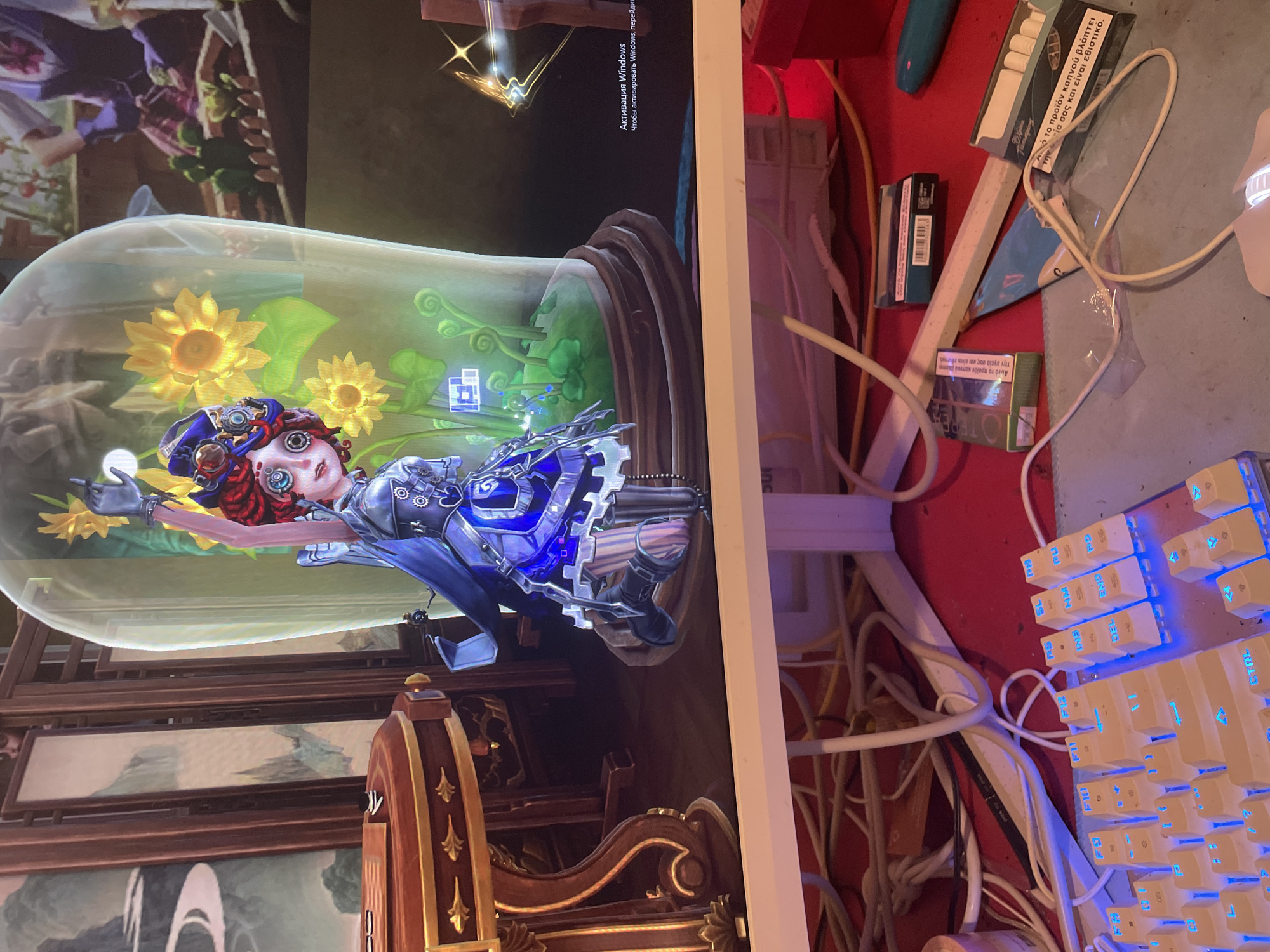 The image displays a computer monitor showing a video game character in a glass terrarium with sunflowers. The monitor is placed on a red surface cluttered with computer wires, a keyboard with illuminated blue keys, and several open packs of cigarettes. The scene is consistent with a home environment, likely in Neoi Epivates, Greece, given the text on the cigarette packs, which is in Greek. There are no people visible in the image.