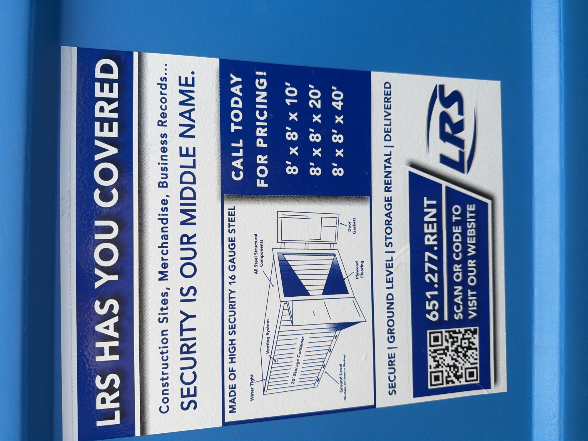 The image shows a close-up view of a bright blue metal surface, likely a storage container or a vehicle, with a white sticker affixed to it. The sticker features a large blue banner at the top with white text that reads "LRS HAS YOU COVERED." Below this, in a smaller font, it states "Construction Sites, Merchandise, Business Records..." followed by a larger, bolder line that says "SECURITY IS OUR MIDDLE NAME."
The middle section of the sticker displays an illustration of a 20' storage container with various components labeled: "Water Tight," "Venting System," "All Steel Structural Components," "Plywood Flooring," "Ground Level (No Steps, Toe Drains or Blocking)," and "Door Gaskets." To the right of the illustration is a blue section with white text indicating "CALL TODAY FOR PRICING!" and a list of container sizes: "8' x 8' x 10'," "8' x 8' x 20'," and "8' x 8' x 40'."
The bottom of the sticker has another blue banner with white text that reads "SECURE | GROUND LEVEL | STORAGE RENTAL | DELIVERED." Below this is a white rectangle containing a QR code, the phone number "651.277.RENT," and text encouraging the viewer to "SCAN QR CODE TO VISIT OUR WEBSITE." To the right of this is the LRS logo in white. The overall impression is an advertisement for storage solutions offered by LRS. The image is well-lit, and the sticker is crisp and clear. There are no people or any environmental context to suggest a specific location within Elk River, United States of America.