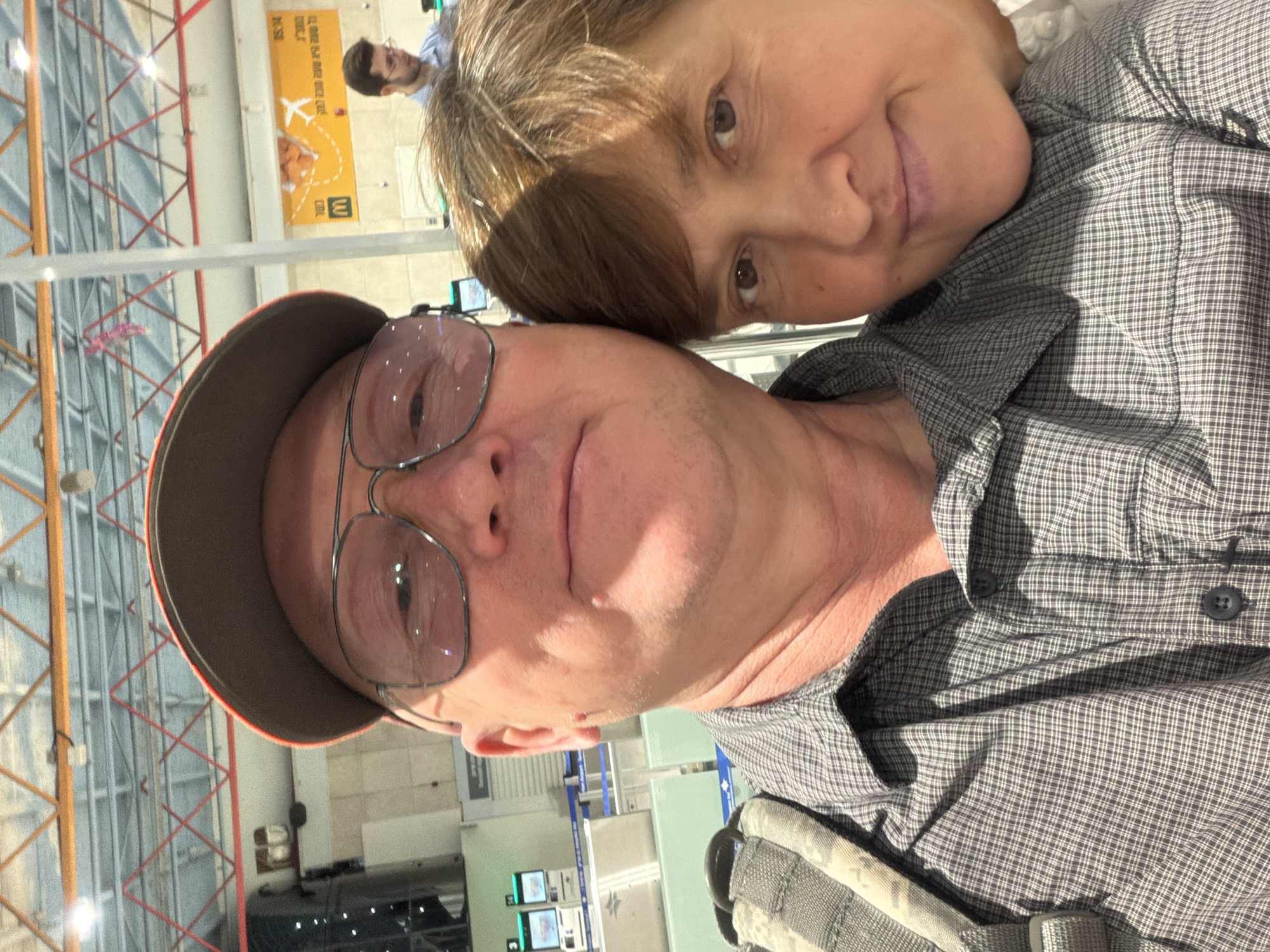 A close-up, selfie-style shot captures two individuals, a man and a woman, at what appears to be an airport in Israel. The man, wearing glasses and a baseball cap, is in the foreground and center-left of the frame. He has fair skin and is dressed in a plaid button-down shirt. The woman is positioned to the right of the man, with her head resting on his shoulder. Her hair is brown and shoulder-length, and she has a gentle smile. Behind them, airport infrastructure is visible, including check-in counters, digital screens, and ceiling beams with red accents. A yellow sign with Hebrew text and a graphic of an airplane is also present, indicating a travel-related location. The overall scene suggests a casual moment captured while traveling.