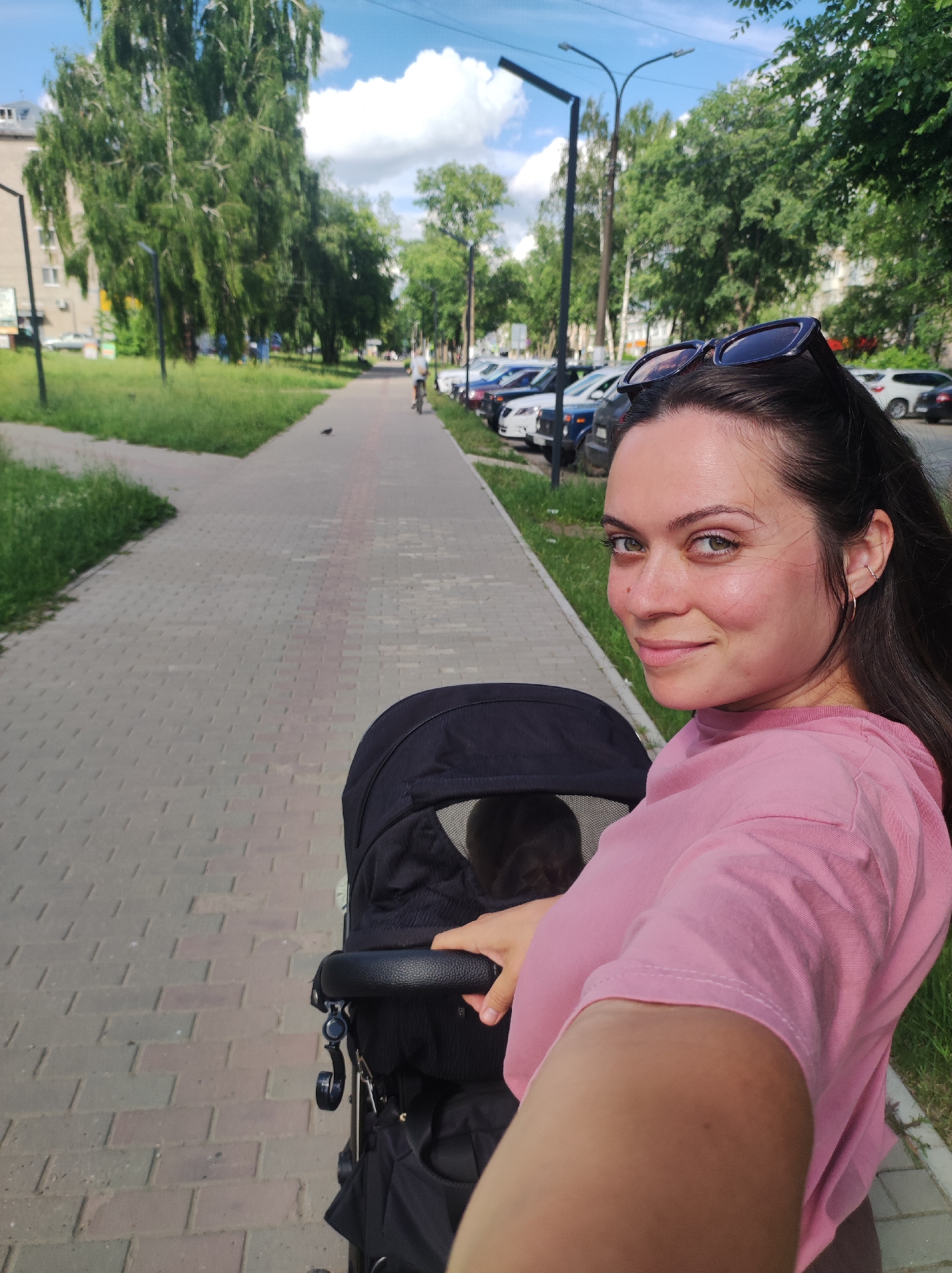 A woman wearing a pink t-shirt and sunglasses on her head is pushing a black stroller down a brick sidewalk. She is looking back at the camera with a slight smile. The stroller has a black canopy, and a baby's head can be seen inside. The woman's hair is dark brown and appears to be blowing in the wind.
The scene is an outdoor setting, likely a park or a residential street, with trees and grass lining the sidewalk. Cars are parked on the right side of the sidewalk. In the background, there is a building and a street with more parked cars. A person on a bicycle can be seen further down the sidewalk.
The time of day appears to be daytime, likely late afternoon given the lighting. The weather seems pleasant, with a blue sky and white clouds. The location is identified as Chaikovsky, Russia. The overall mood is casual and relaxed, suggesting a leisurely walk. No visible text is present in the image.
