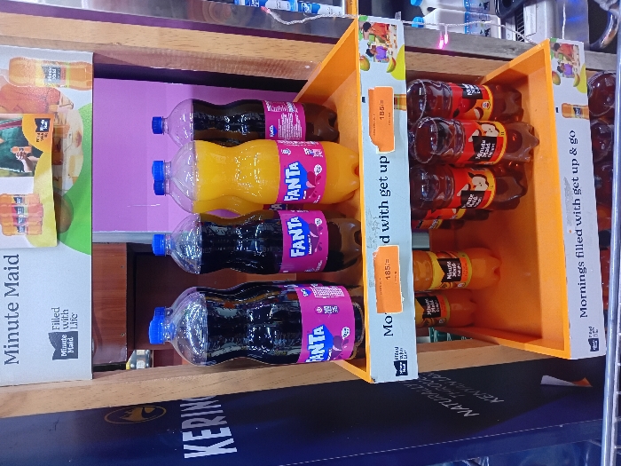The image displays an indoor retail beverage display stand located in Naro Moru, Kenya. The stand features two main orange-colored tiered shelves constructed of wood and plastic.
On the upper shelf, five 1.25-liter plastic soda bottles are arranged: three bottles contain a dark-colored carbonated liquid (Fanta Blackcurrant), and two bottles contain an orange-colored carbonated liquid (Fanta Orange). All bottles are sealed with blue plastic caps. Two orange price labels affixed to the shelf front, partially obscured, indicate a price of "185/=". Above this shelf, a white sign with "Minute Maid" branding is visible, featuring product images and text such as "Filled with Life."
The lower shelf holds smaller plastic bottles of Minute Maid juice. Two bottles containing an orange-colored liquid (likely orange juice) are visible on the left side of this shelf, alongside a row of four dark reddish-brown bottles with apple graphics (Minute Maid Apple Juice) on the right. The front edge of this shelf displays the slogan "Mornings filled with get up & go."
To the immediate left of the display stand, a dark blue banner with partial text "KERING..." and "NATURALLY THROUGH KENYAN..." is visible. A light purple wall forms the background directly behind the display. A commercial refrigeration unit with white bottles is partially visible on the far right background. The chrome handle of a shopping cart is visible in the foreground at the bottom of the image. No individuals are present within the scene.