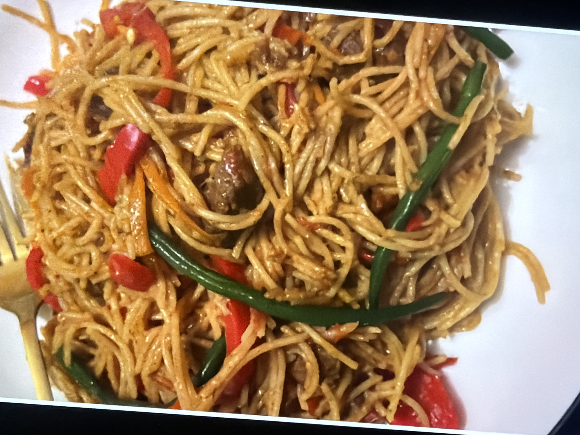 A plate of Pad Thai noodles is presented from a slightly elevated, angled perspective, with a wooden fork resting on the upper left portion of the dish. The noodles, a light brown color, are interwoven with vibrant red bell pepper slices and dark green string beans. Small pieces of meat are also visible throughout the dish, suggesting it contains beef or chicken. The background is a plain white surface, likely a plate. The overall lighting is even, highlighting the textures and colors of the food. The scene is devoid of any human subjects or environmental context beyond the food itself.