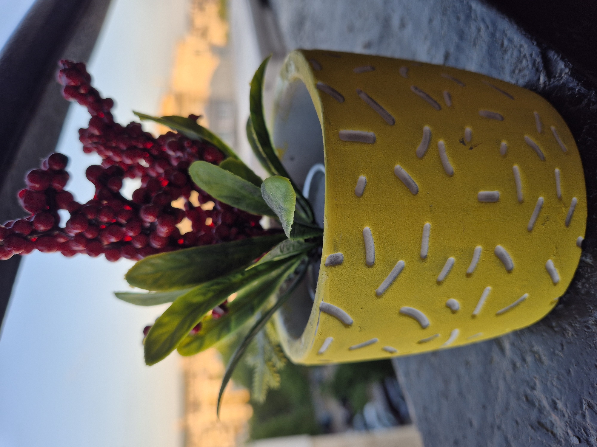 A close-up, low-angle shot focuses on a yellow, textured ceramic pot with a decorative, dashed pattern. Inside the pot, artificial greenery with a sprig of red berries emerges. The pot sits on a dark, textured surface, possibly a balcony railing, with a blurred urban background visible under a clear sky.