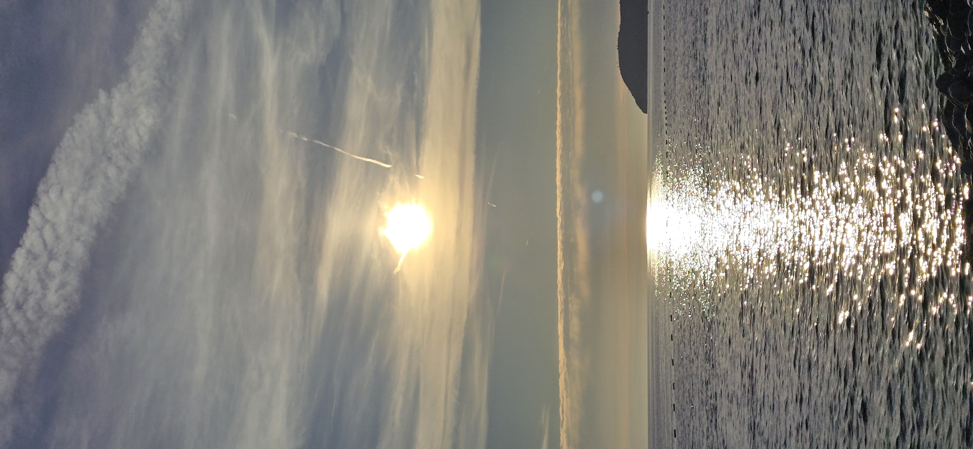 The image captures a coastal landscape in Bar, Montenegro, during late afternoon or sunset. The sky, transitioning from pale blue at the top to a warm yellow-orange closer to the horizon, features scattered, wispy clouds and multiple contrails. The sun, a brilliant white-yellow sphere emitting prominent light rays and lens flare, is positioned slightly left of the image's vertical center. A distinct, diagonal contrail extends from the upper right, while a fainter, shorter contrail is visible below the sun. A horizontal band of denser clouds lies just above the sea horizon. The lower portion of the image displays the sea with a gently rippled surface. A luminous path of sun glint, appearing as countless sparkling points of light, extends from the horizon towards the foreground. Approximately eight small, dark, rectangular buoys are arranged in a horizontal line across the mid-left to mid-center of the water. On the far right, a dark, rounded landmass or island is discernible against the horizon. The immediate foreground on the bottom right reveals several dark, irregular rocks along the water's edge. No human figures or other significant man-made objects are present in the scene.