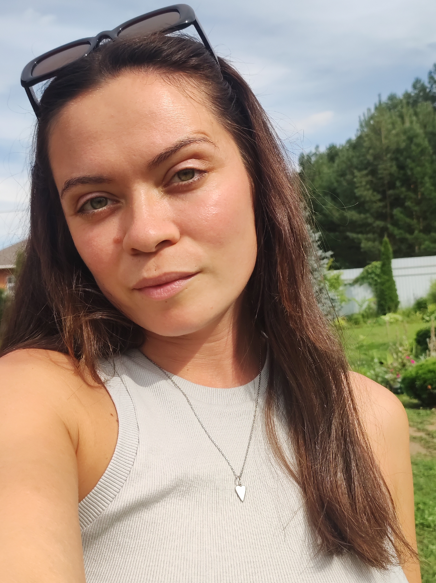 In a medium, slightly low-angle, eye-level shot, a young woman with long, dark brown hair and light green eyes is captured in a selfie. Sunglasses are perched on top of her head. She wears a light gray, ribbed tank top with a thin silver chain and a heart-shaped pendant. The background suggests a sunny day with a clear blue sky dotted with white clouds and a line of green trees. A white fence is visible, and the setting appears to be a residential backyard, possibly in the city of Фоки, Russia, judging by the architecture and vegetation. The lighting indicates it's daytime.