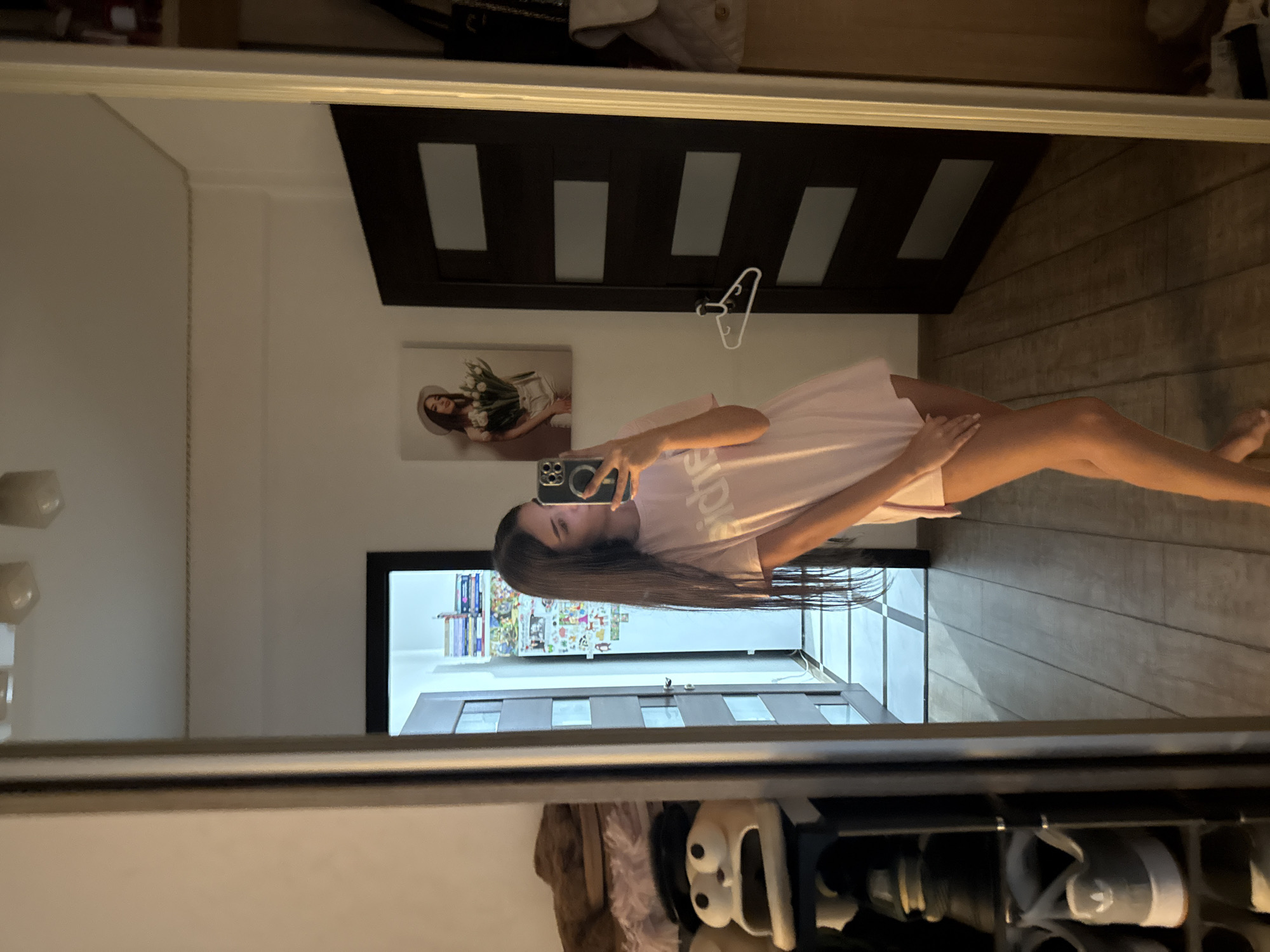 A young woman with long dark hair is taking a mirror selfie in what appears to be a bedroom or hallway in Chișinău, Moldova. She is wearing a light pink, oversized t-shirt that extends past her mid-thigh. The mirror she is using is large and reflects a significant portion of the room. To her left, a shoe rack is visible, holding several pairs of shoes, including a distinctive pair of novelty slippers with large cartoon eyes and an open mouth. Further to the left, a furry brown object, possibly a blanket or garment, is draped over something.
In the reflection of the mirror, behind the woman and to her right, a dark-colored door with five rectangular frosted glass panels is visible. A white hanger is hanging on the door. Above the door, a framed picture is hung on the wall, depicting a woman wearing a hat and holding a bouquet of white tulips. The wall itself is white. The floor in the reflection is made of light-colored wood planks, with a checkered pattern of lighter and darker tiles in the hallway beyond.
To the far right of the reflection, part of a wardrobe or closet is visible, with a white backpack or purse hanging on it. The lighting in the room appears to be artificial, with ceiling lights casting some shadows. The overall environment suggests a domestic setting.