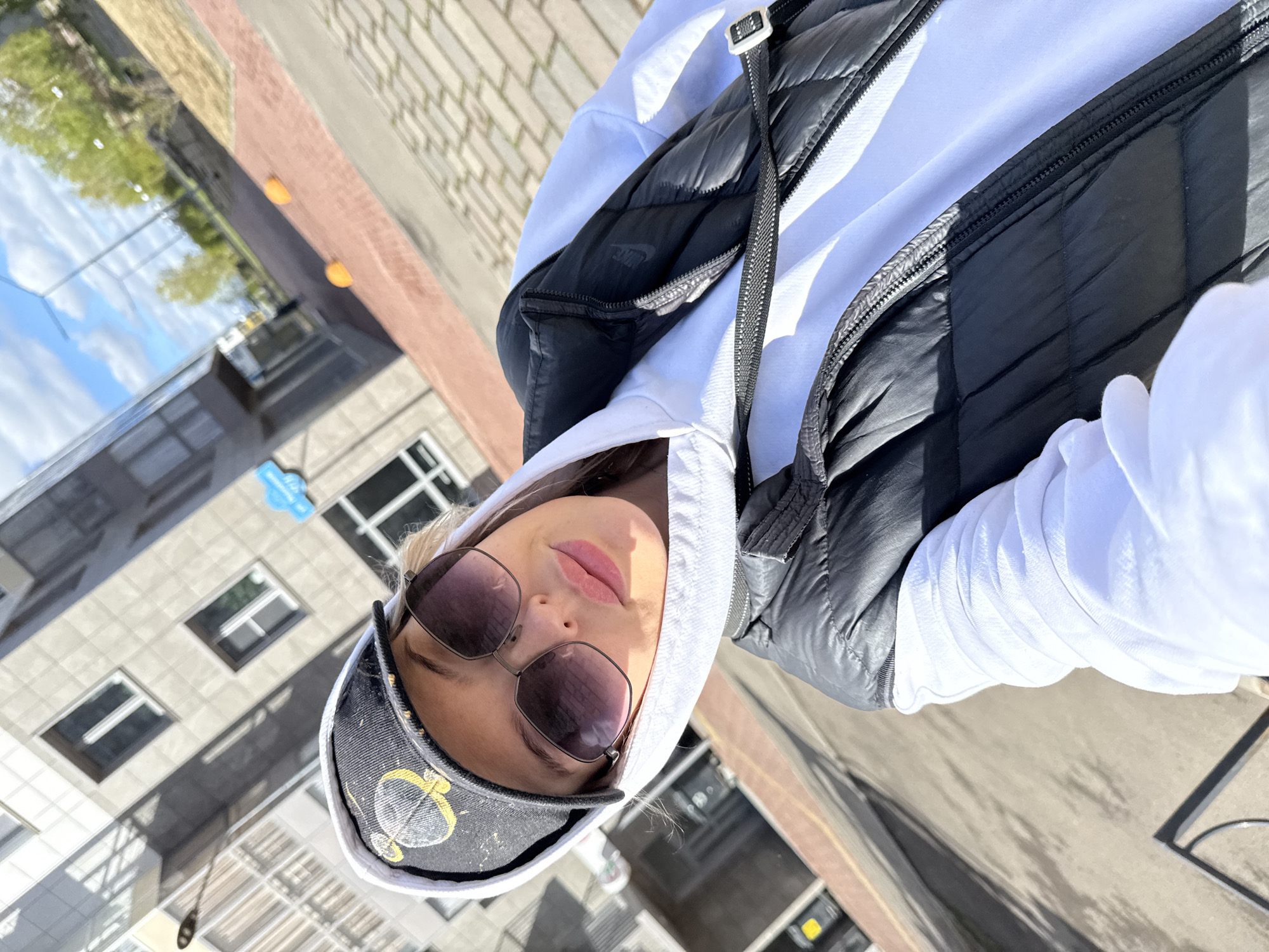 A young woman takes a selfie in Krasnoyarsk, Russia. She wears sunglasses, a decorated cap, a white hoodie, and a black vest. The building and street behind her are reflected in the glass. The sky is blue with some clouds. The image is slightly rotated counterclockwise.