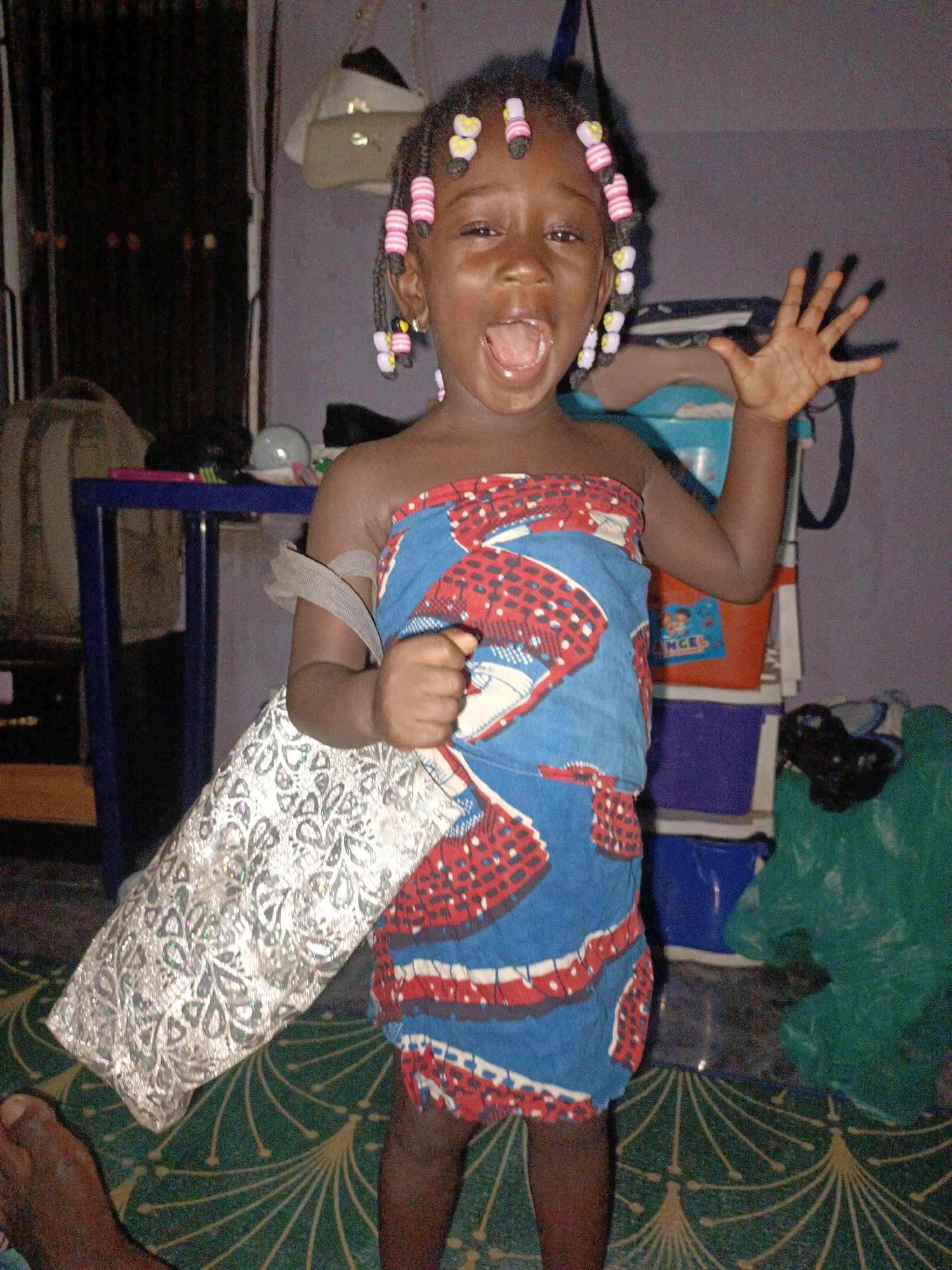 A young, dark-skinned child stands in an indoor setting, possibly a home, in Akwanga, Nigeria. The child has dark hair adorned with pink and yellow beads. She is wearing a brightly colored blue, white, and red patterned fabric wrapped around her chest. The child has her mouth open and one arm raised in the air with an open palm, while the other hand holds a large, silver-patterned bag. The background includes a table with miscellaneous items, stacked colorful plastic storage containers, and a green plastic bag. The floor has a dark green pattern on it. A foot is partially visible in the bottom left corner of the frame.