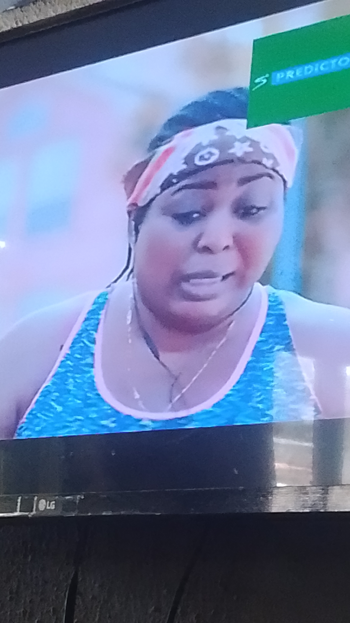 A medium shot captures a woman with dark skin, wearing a pink and white patterned bandana and a blue tank top with pink trim. Her eyes are looking slightly down and to the left, and her mouth is partially open as if speaking. She is wearing a delicate silver chain around her neck. The background is softly blurred, with a green rectangular logo in the upper right corner that reads "PREDICTOR." The bottom of the frame shows the black frame of a television, with the "LG" logo visible on the bottom left. This image was taken in Lokoja, Nigeria.