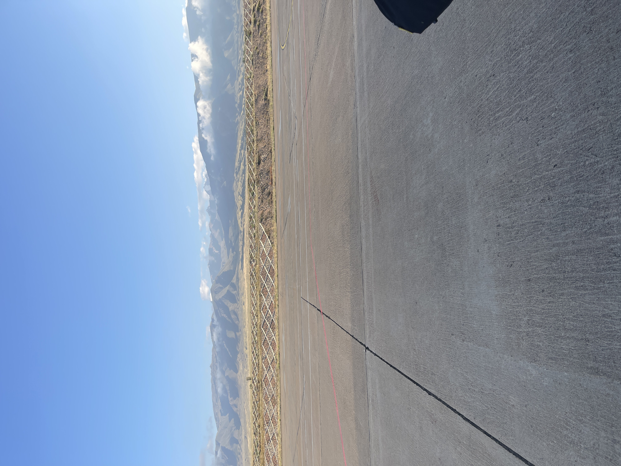 The image displays an outdoor scene, likely an airport tarmac, with a concrete surface in the foreground marked with lines, including a red line and a yellow curved line. In the middle ground, there is a tiered hillside with a diamond-shaped fence structure, possibly a barrier or a retaining wall, filled with rocks. The background features a mountain range under a clear blue sky with some clouds. The overall impression is that of a vast, open space with a natural landscape in the distance. The lighting suggests it is daytime, possibly morning or late afternoon. There are no people or animals visible in the image. The location is identified as being in the city of Choktal, Kyrgyzstan.