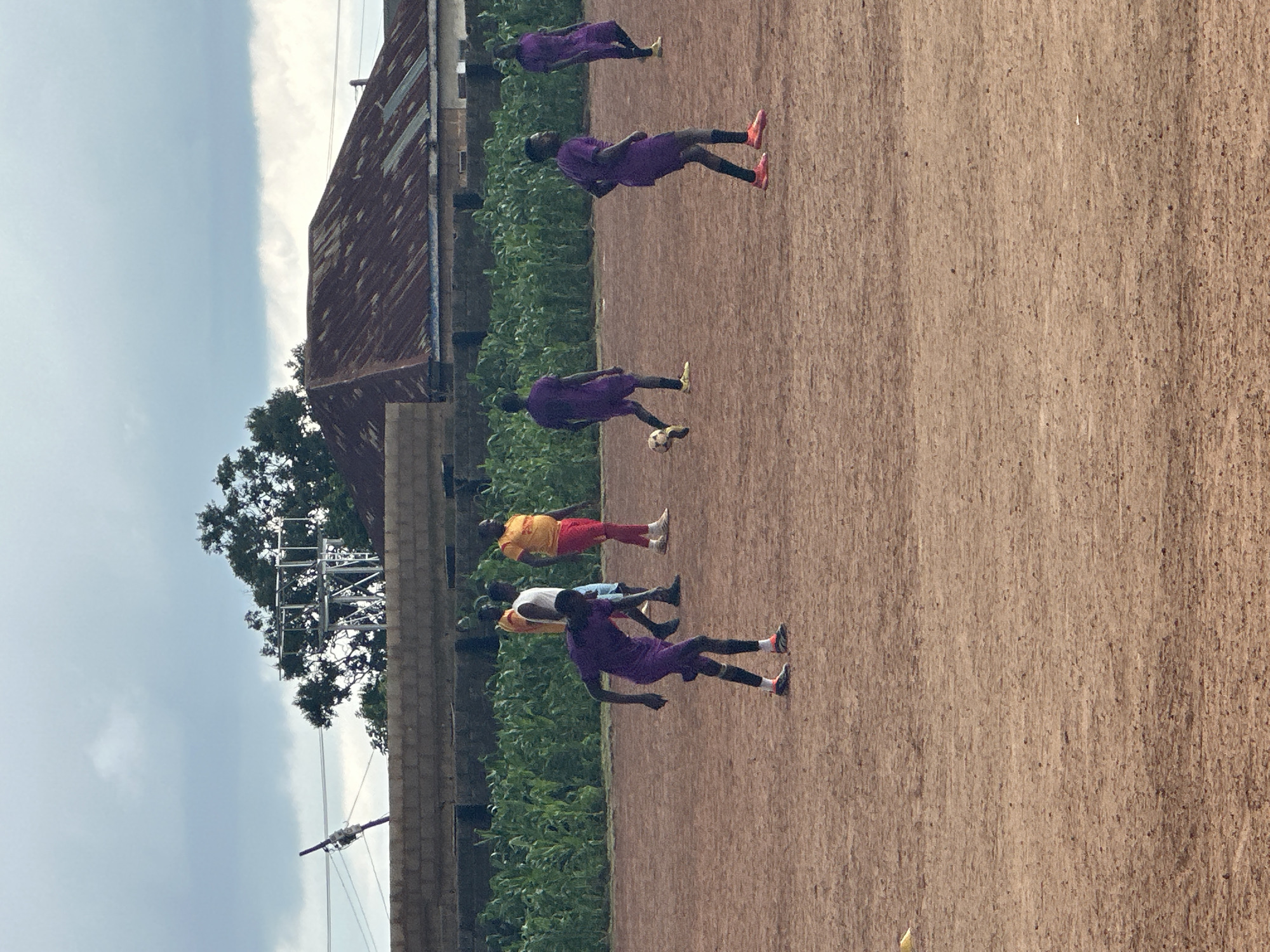 The image depicts an outdoor scene, likely a soccer field, in Rafin Bauna, Nigeria. Several individuals are present, dressed in athletic wear. Some are wearing purple uniforms, while others have on yellow shirts with red and blue accents. One person in a purple uniform is actively engaged with a soccer ball, appearing to be in the process of kicking or controlling it. Other individuals are standing or walking on the dirt-colored field. In the background, a brick wall runs horizontally, beyond which is a dense row of green vegetation, possibly corn crops. Further in the distance, a building with a corrugated metal roof is visible, along with trees and a utility pole with electrical wires. The sky is overcast with clouds.